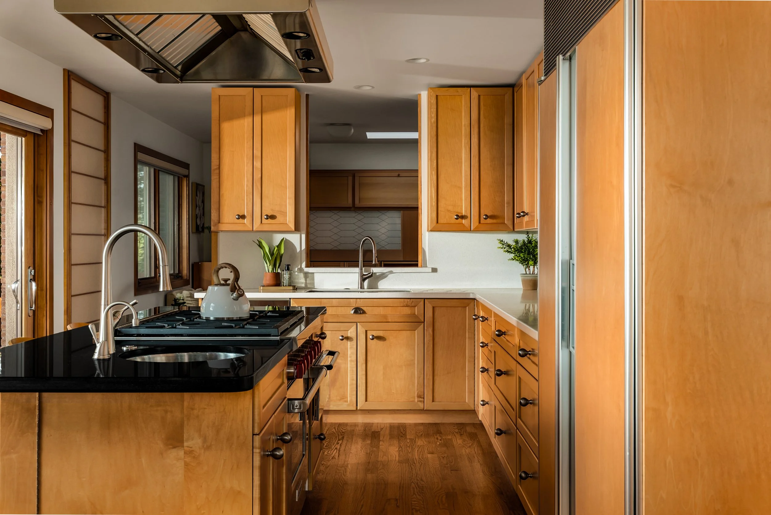A kitchen with wooden cabinets, a black stove with a white kettle, and a sink with a plant and soap dispenser on the counter.