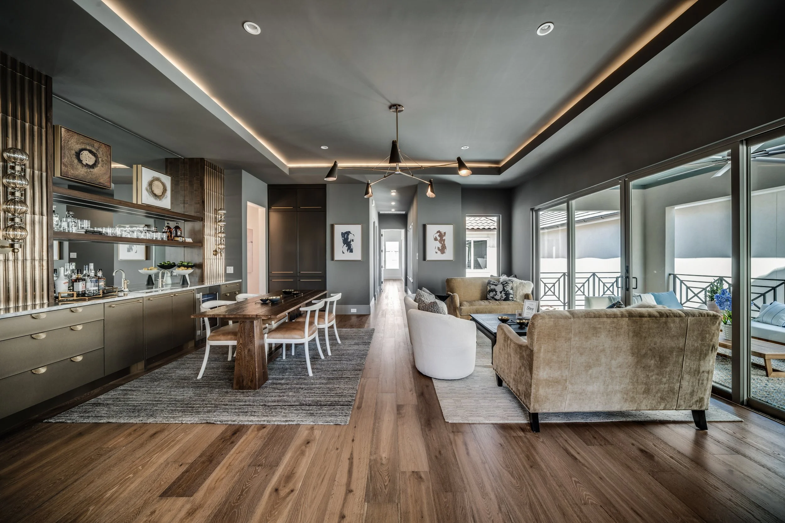 Modern living room with hardwood floors, a gray ceiling with recessed lighting, large windows with sliding doors, a hanging light fixture, a beige sofa with patterned cushions, a wooden dining table with white chairs, and wall art.