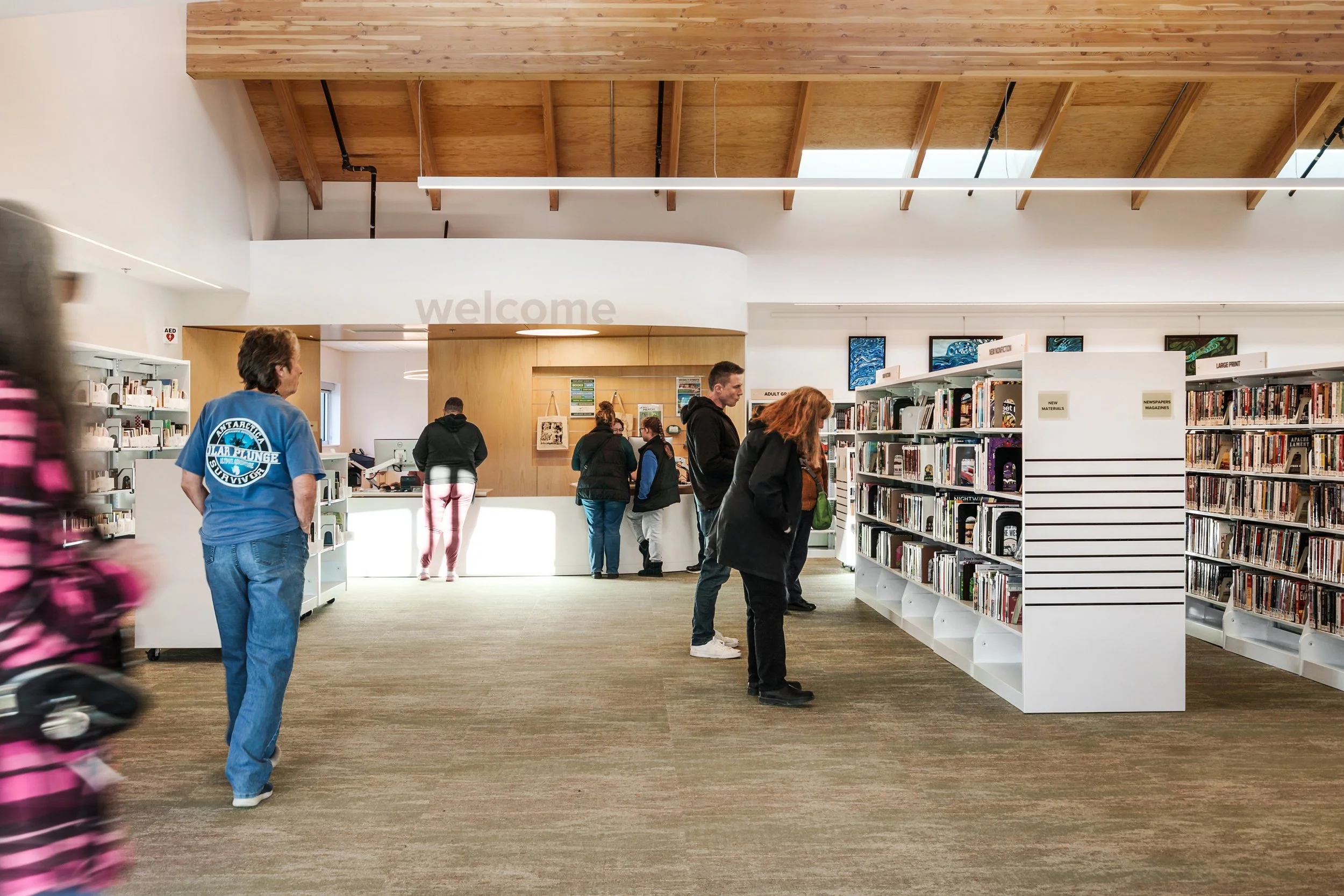 People browsing bookshelves in a modern library with a high wooden ceiling and white walls.