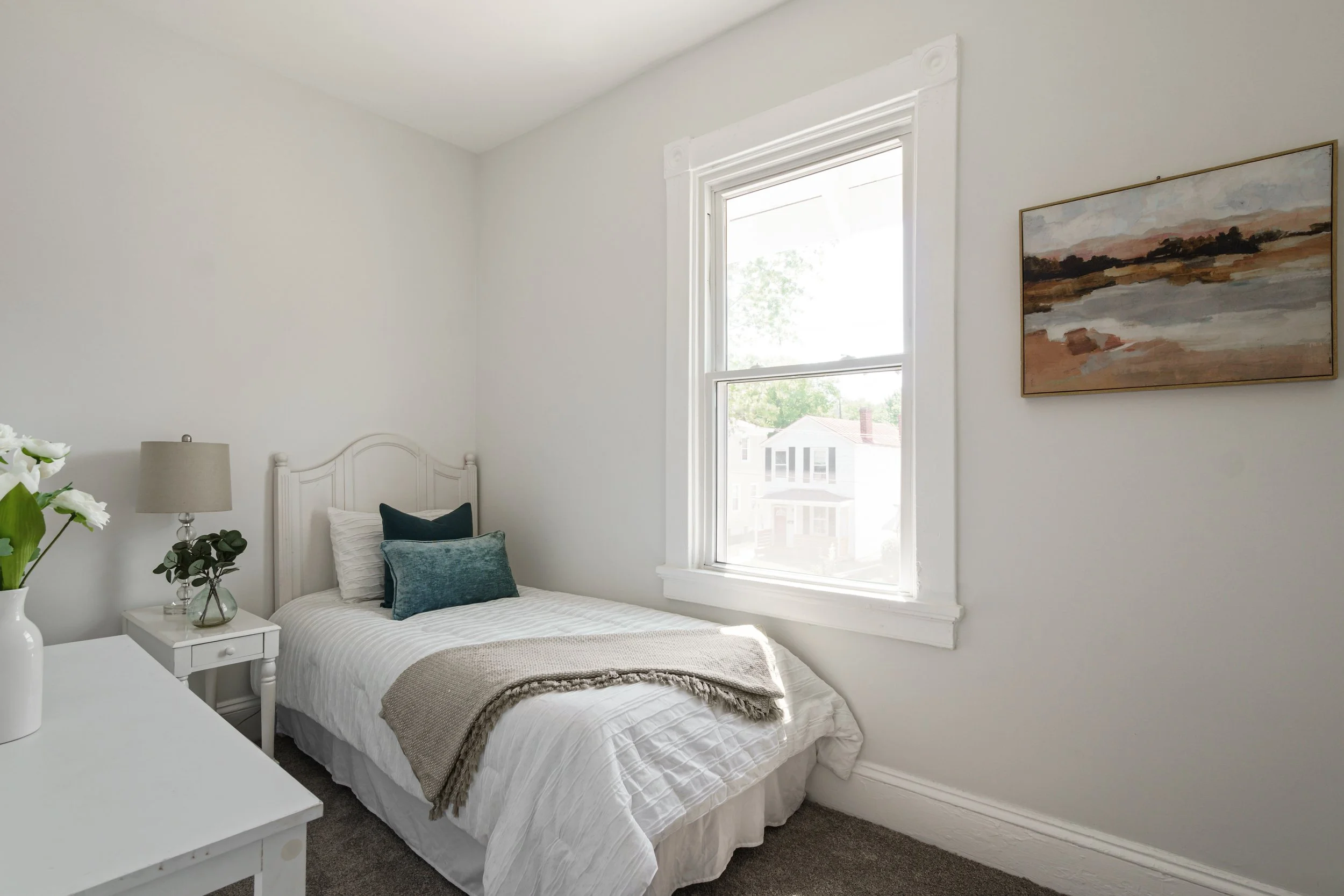 A bright, minimalist bedroom with white walls, a bed with white bedding and blue pillows, a small white nightstand with a lamp, a potted plant, and a framed landscape painting hanging on the wall. There is a large window allowing natural light into t
