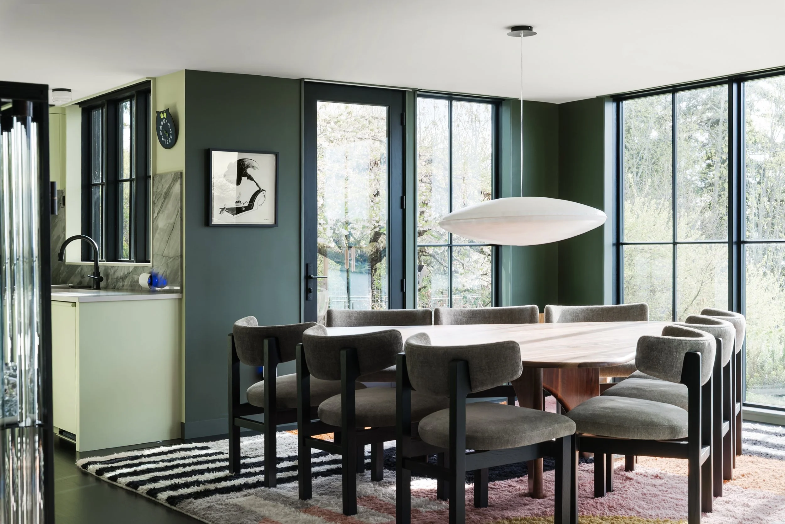 Modern dining room with a large oval wooden table, upholstered chairs, large windows with a view of trees, a white oval hanging light fixture, and framed artwork on the wall.