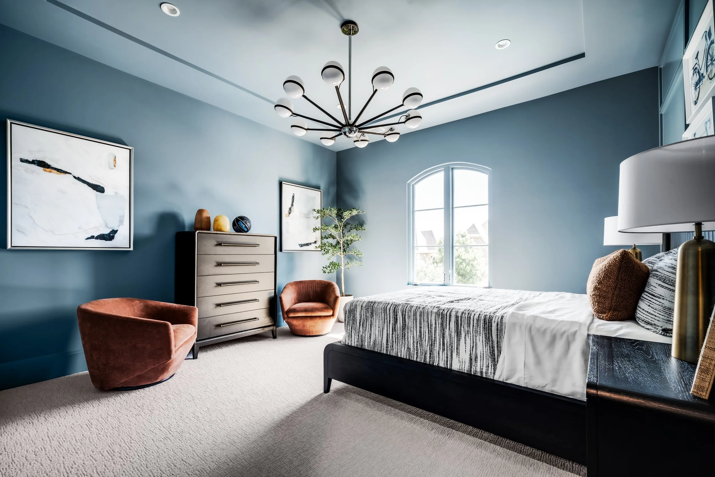 A cozy bedroom with blue walls, a large window, a bed with striped bedding, a pair of orange velvet chairs, a grey dresser with vases, and modern wall art, illuminated by a contemporary chandelier.