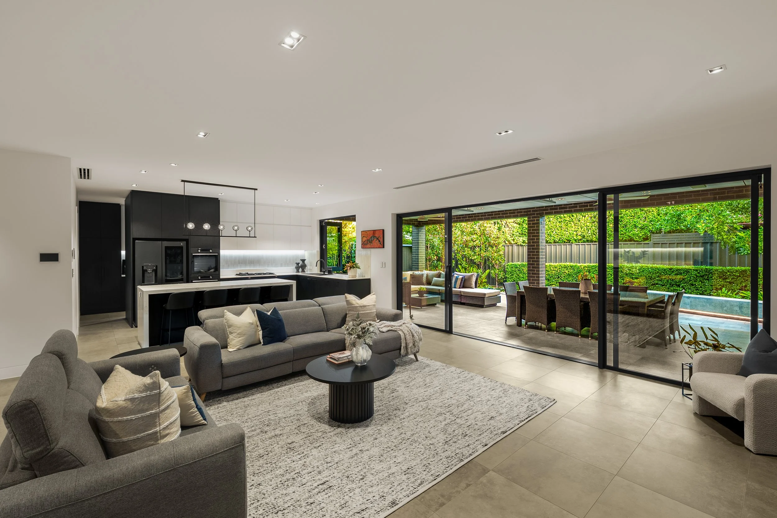 Open-plan living room with gray sofas, decorative pillows, black coffee table, and large sliding glass doors leading to backyard with outdoor seating and greenery.
