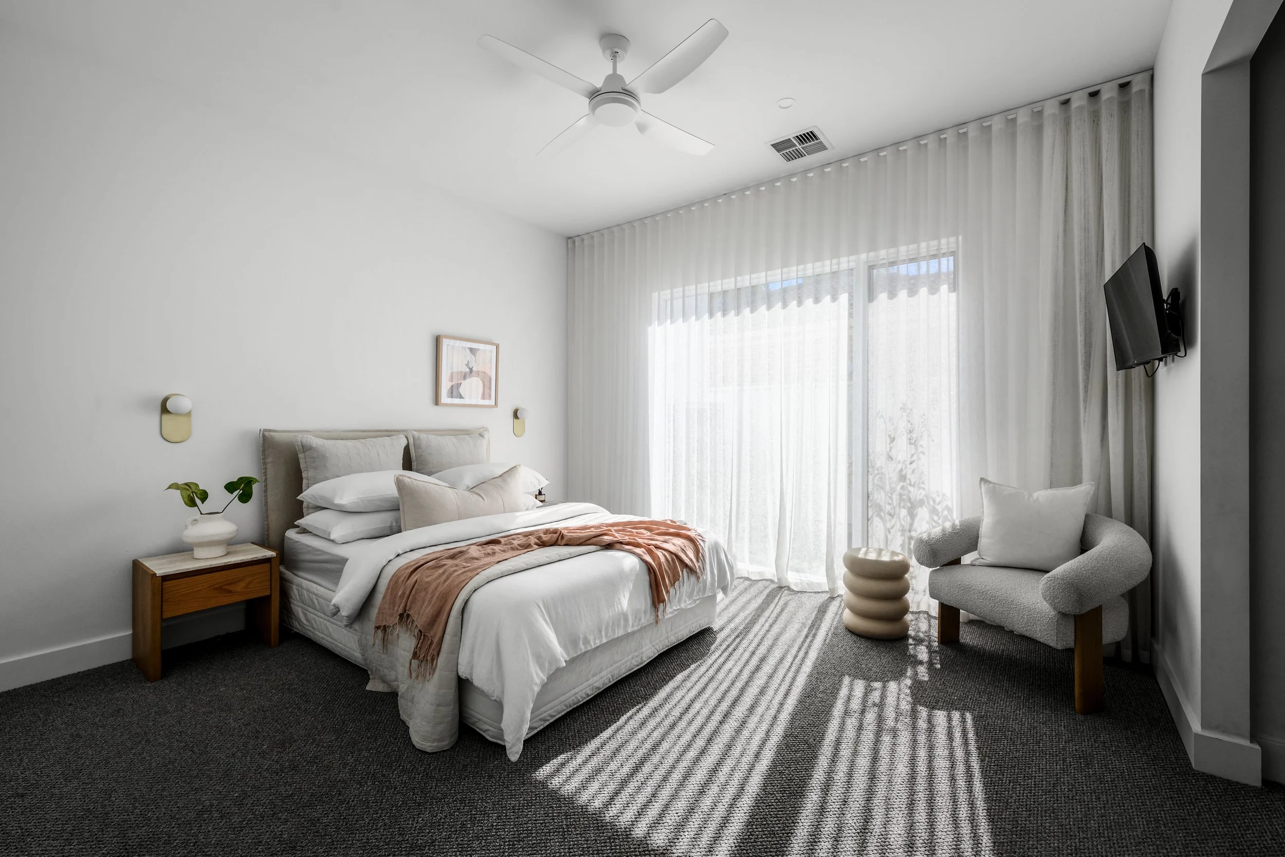 A modern bedroom with a neatly made bed, a bedside table with a plant, abstract wall art, a white ceiling fan, sheer curtains, a small armchair, and a wall-mounted TV.