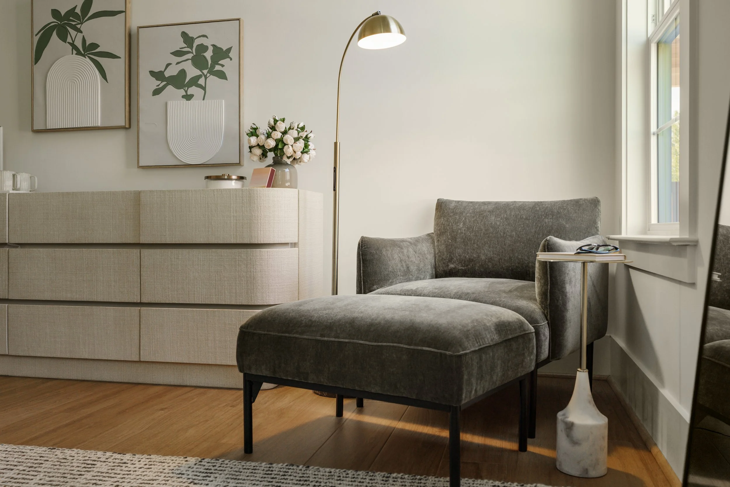 A cozy living room corner with a dark gray upholstered chair, side table, and a tall gold floor lamp. There is a large window with white trim letting in natural light, and a wall with minimalist framed art and a vase of white roses on a nearby surfac