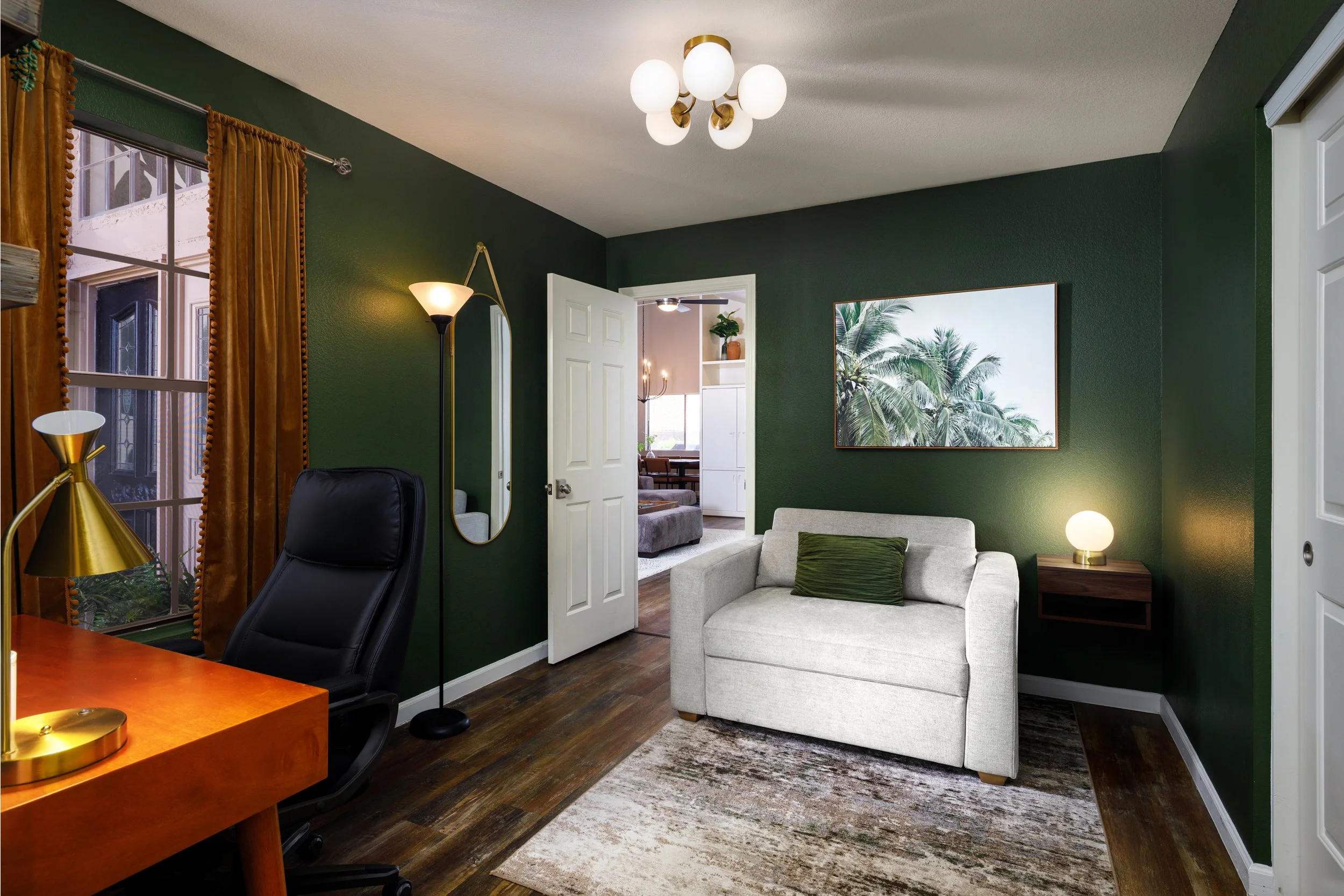 Living room with dark green walls, white ceiling, and wooden flooring. There are gold and white curtains on a window, a small desk with a black office chair, and a black floor lamp. A white sofa with green pillows is next to a wooden side table with 