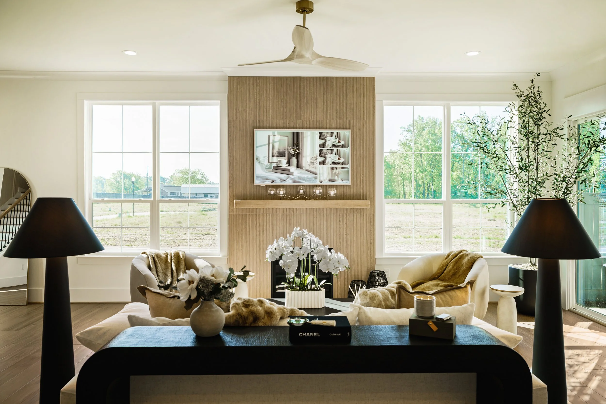 Living room with large windows, beige furniture, a fireplace with a gold decorative piece on top, a TV mounted above it, ceiling fan, black table lamps, and indoor plants.