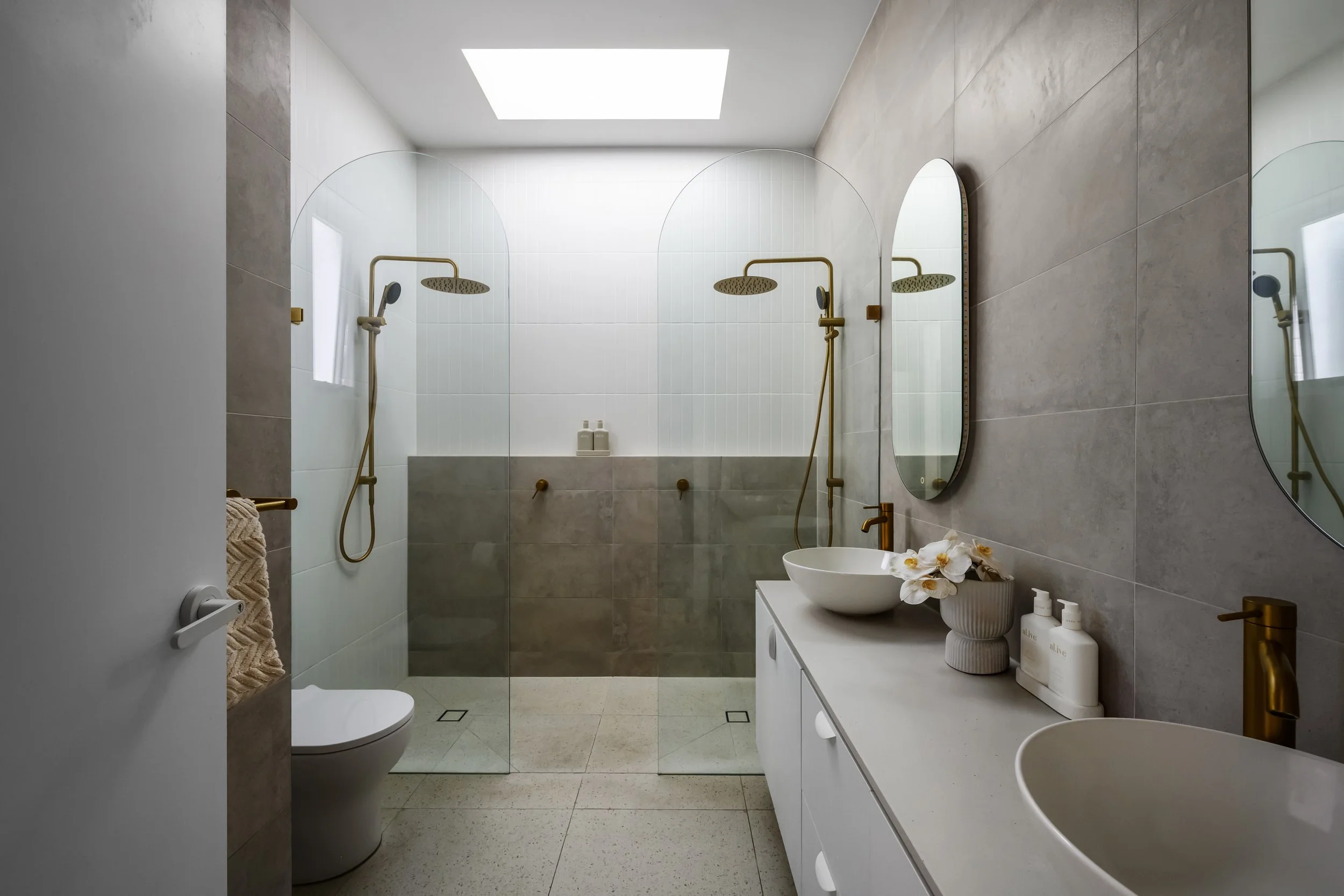 Modern bathroom with two walk-in showers, a toilet, a white vanity with a vessel sink, and decorative flowers. The space has neutral tones, minimalistic design, large mirror, and a skylight.