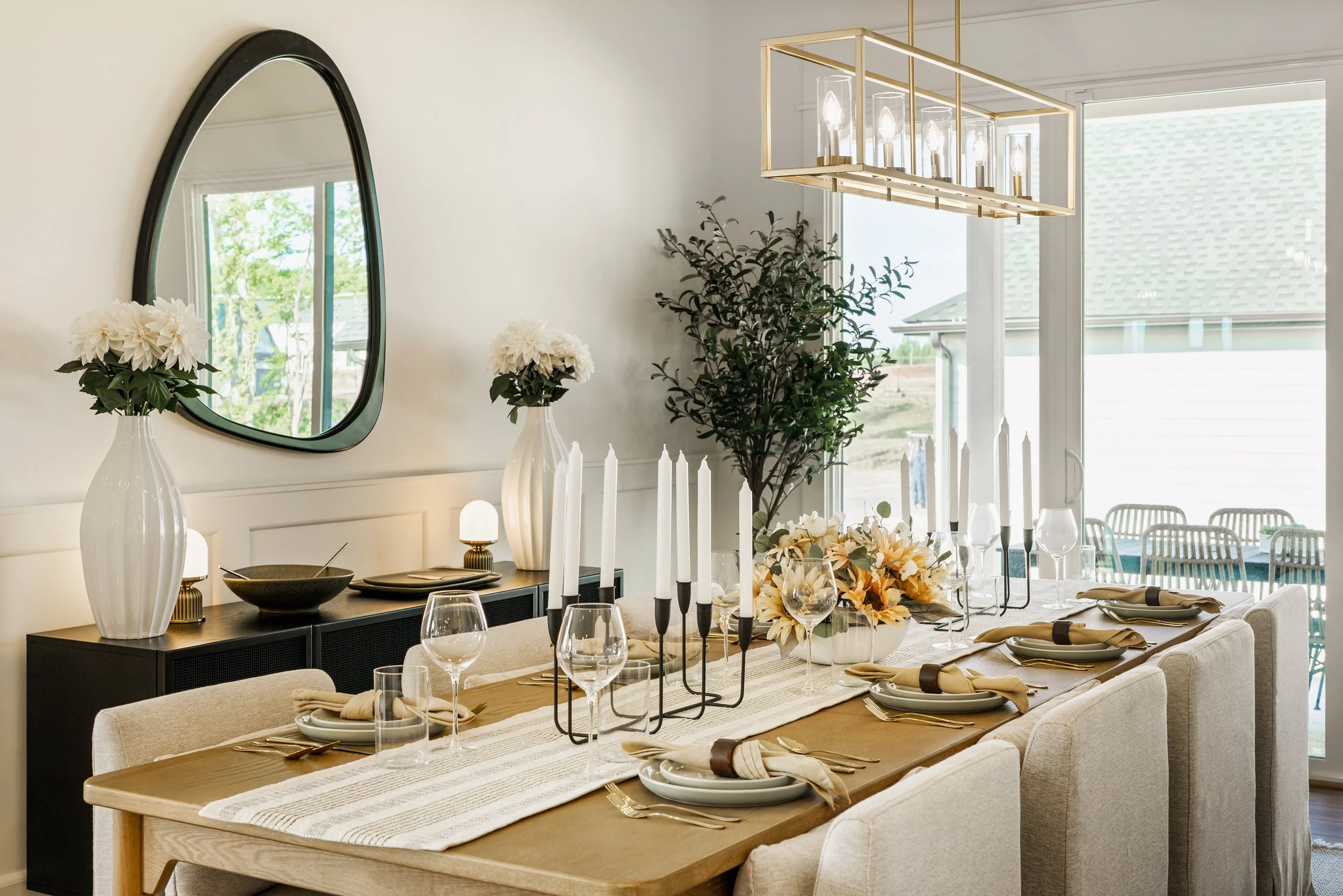 A dining room with a wooden table set for a meal, with beige upholstered chairs, gold and black candlesticks, and a floral centerpiece. White vases with white flowers sit on a black sideboard beneath a large oval mirror. A modern chandelier with expo