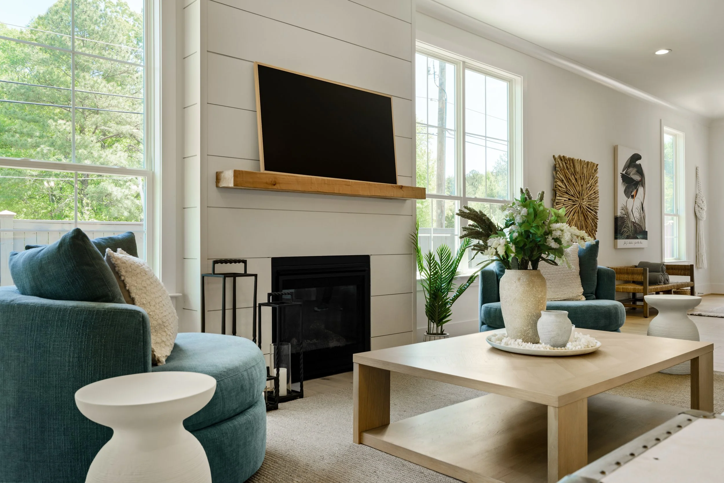 Living room with a teal sofa, white side tables, a wooden coffee table, and large windows with a view of green trees outside. There is a modern fireplace, a mounted flat-screen TV above a wooden mantel, and various decorative elements including plant