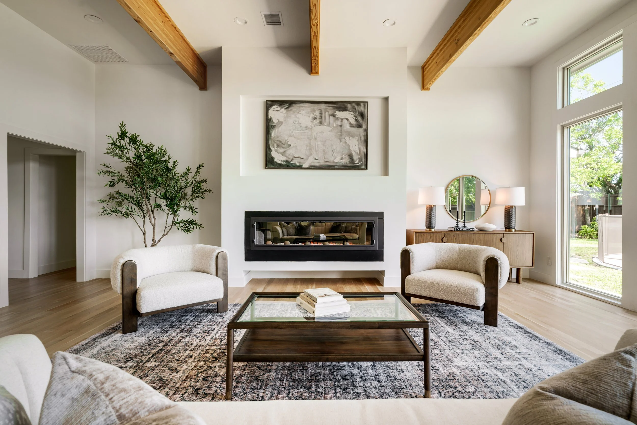 Living room with white walls, large windows, a fireplace, beige armchairs, a wooden coffee table with books, a patterned rug, two table lamps, a round mirror, indoor plants, and wooden ceiling beams.