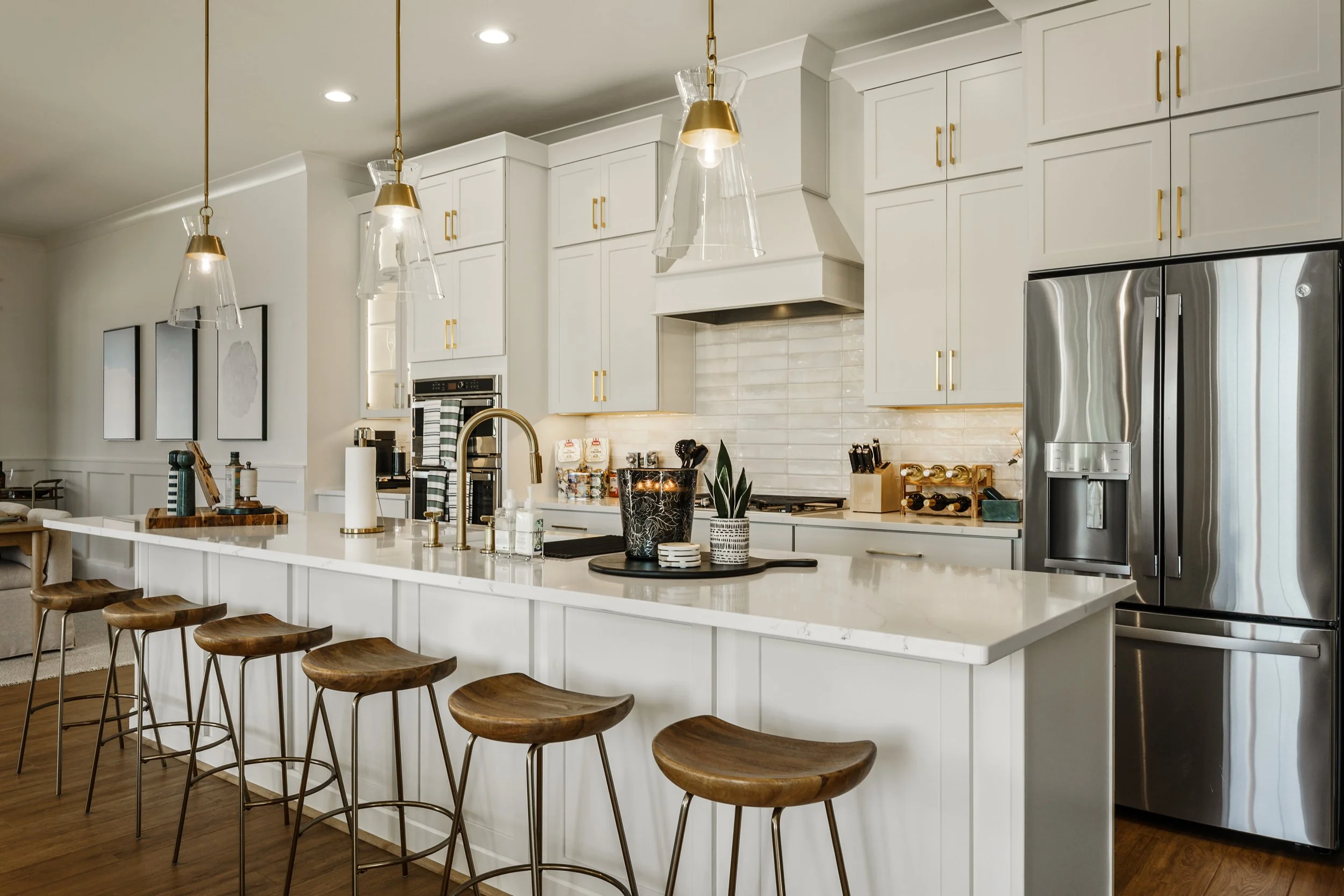Modern white kitchen with gold handles, wood barstools at a large white island, pendant lights, stainless steel refrigerator, and decorative vases on the countertop.