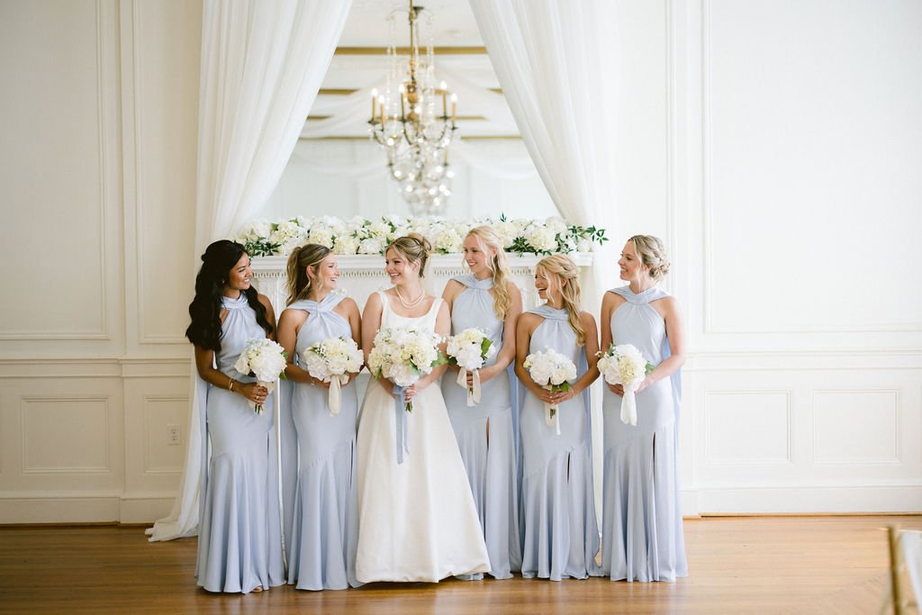 A bride and five bridesmaids standing together in a bright, elegant room with white walls and curtains, each holding a bouquet of white flowers.