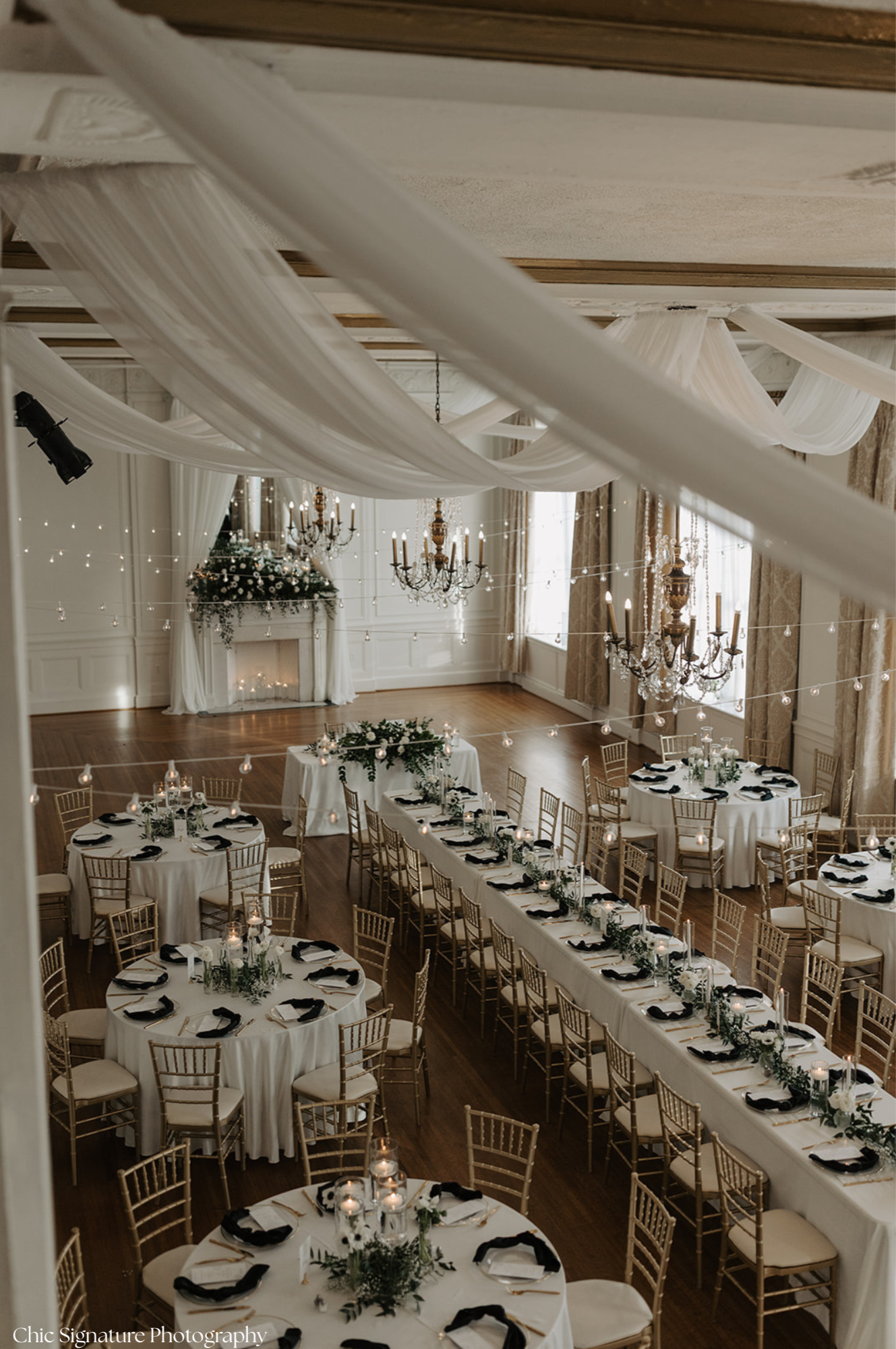 A wedding reception hall decorated with white drapes, chandeliers, string lights, floral arrangements, and round tables with black napkins, candles, and gold chairs.