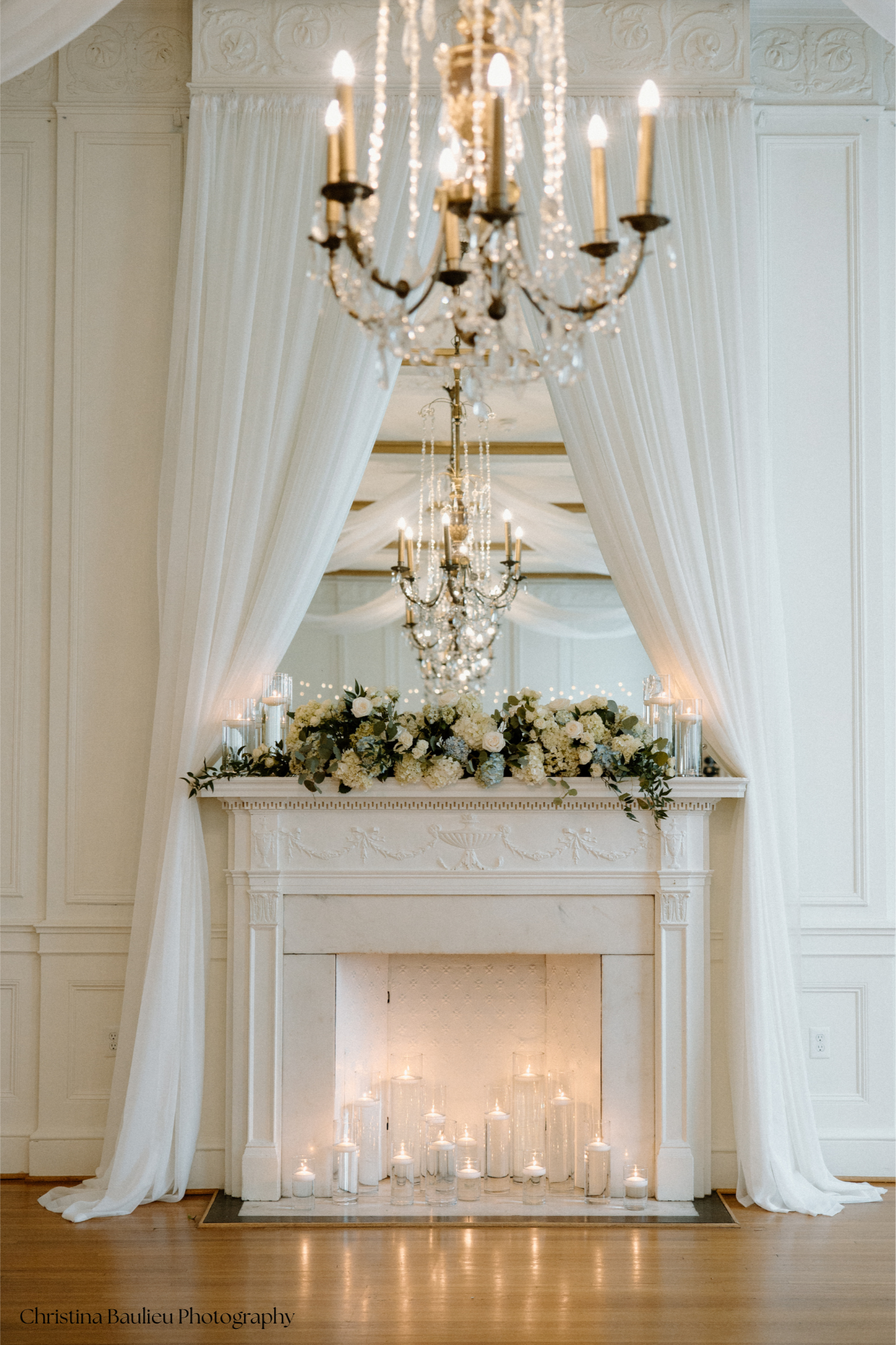 Elegant interior with a white fireplace mantel decorated with a floral arrangement of white and green flowers and greenery. The mantle is in front of a large mirror and surrounded by white drapes tied back, revealing a grand room. Five candles in gla