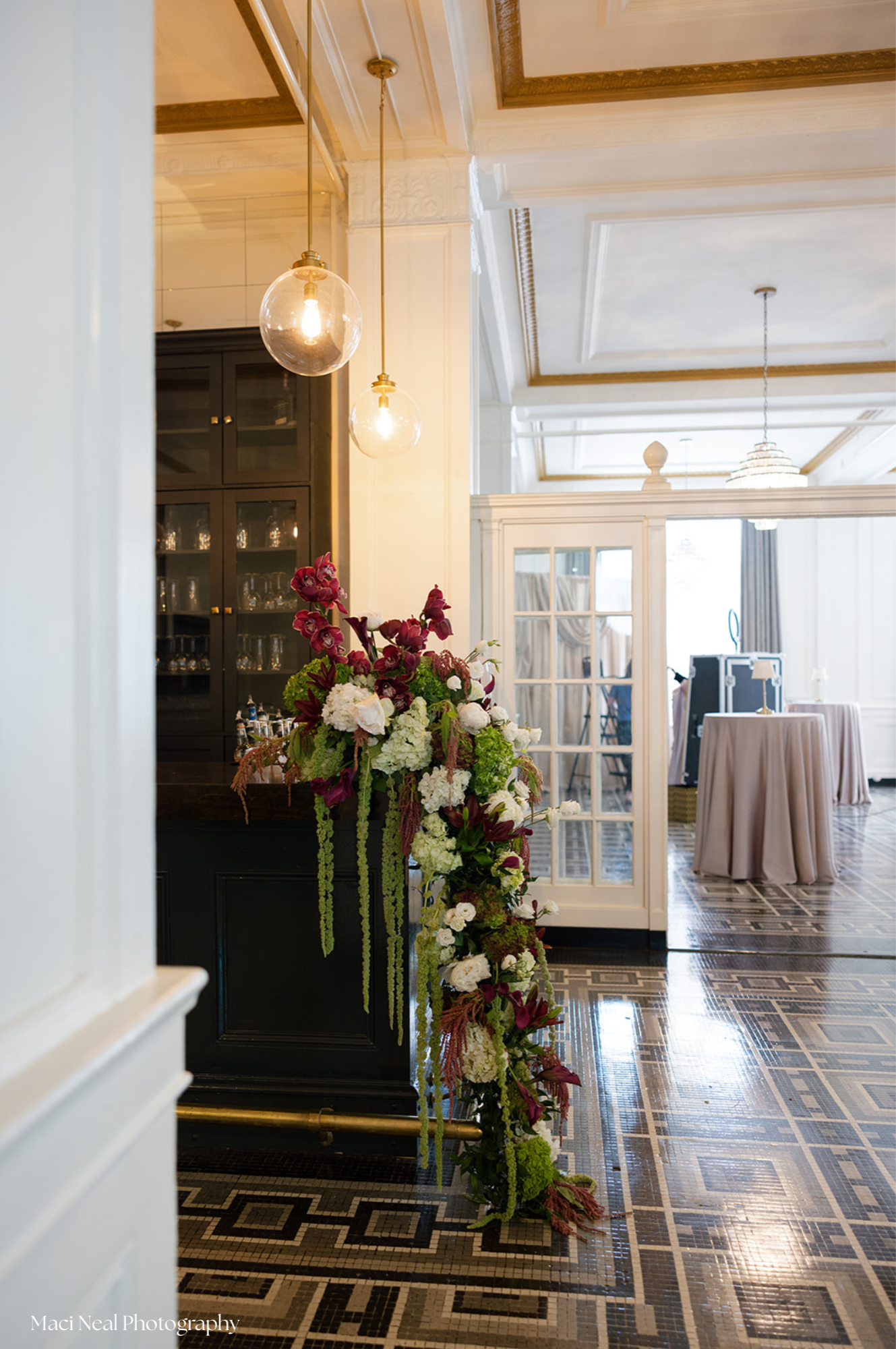 Elegant room with pendant lights, a floral arrangement on a black cabinet, and a white-paneled ceiling with gold trim. In the background, there are tables with pink tablecloths and a chandelier.