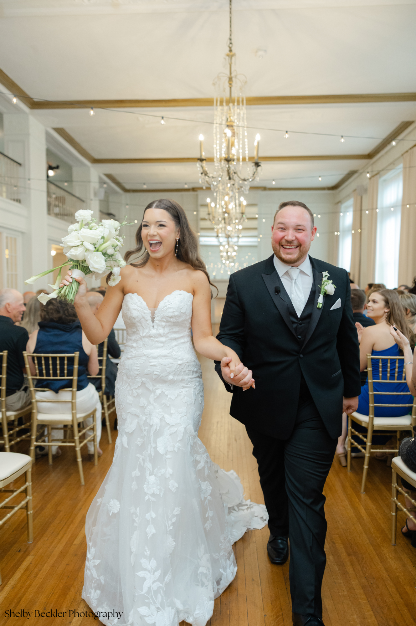 A newlywed couple walking down the aisle after their wedding ceremony, smiling and holding hands, in a decorated wedding venue with chandeliers and seated guests.