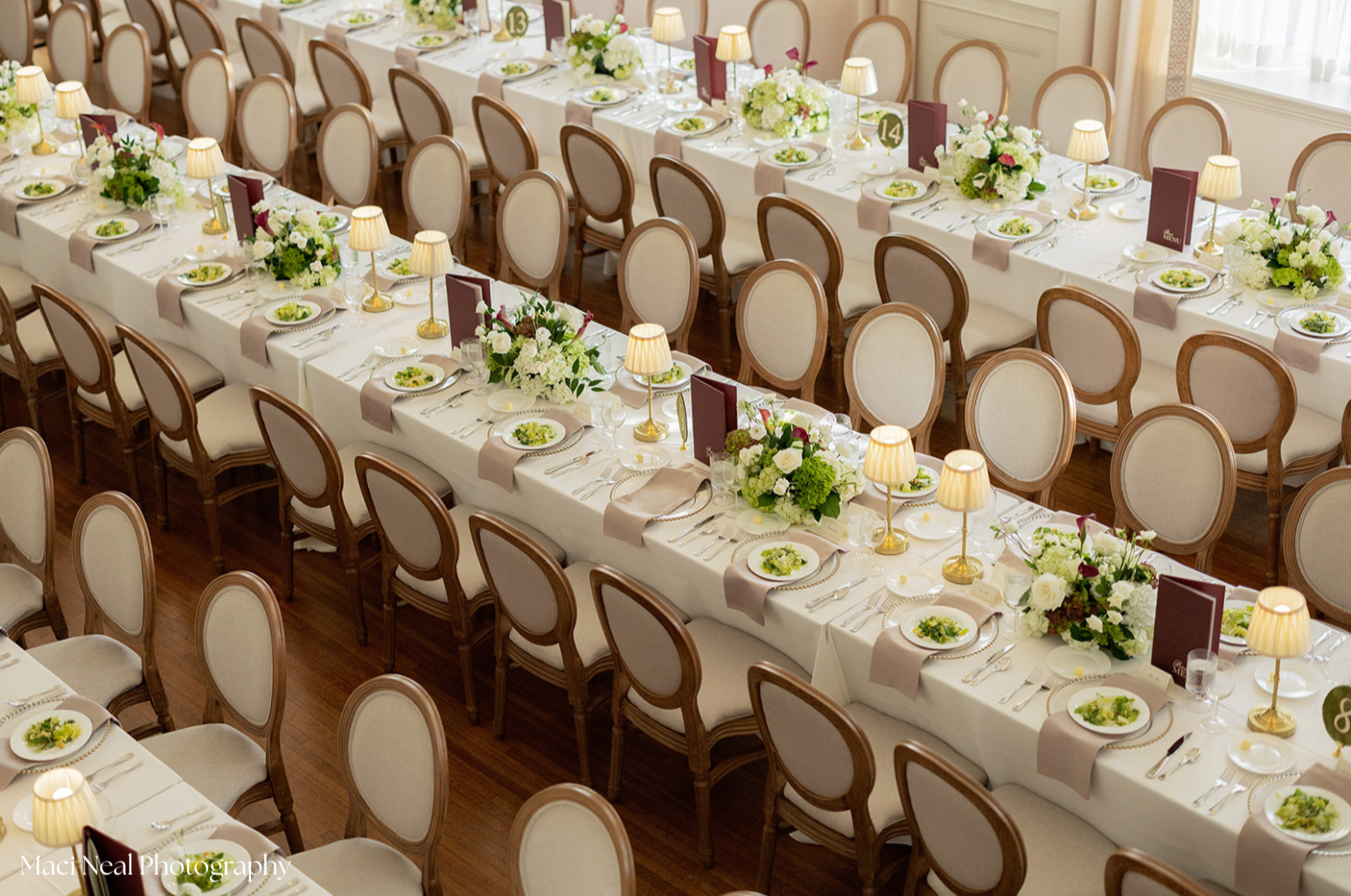 Elegant banquet hall with long tables set with white tablecloths, floral centerpieces, small table lamps, and place settings including plates with salad, cutlery, glasses, and menu cards, ready for a formal event.
