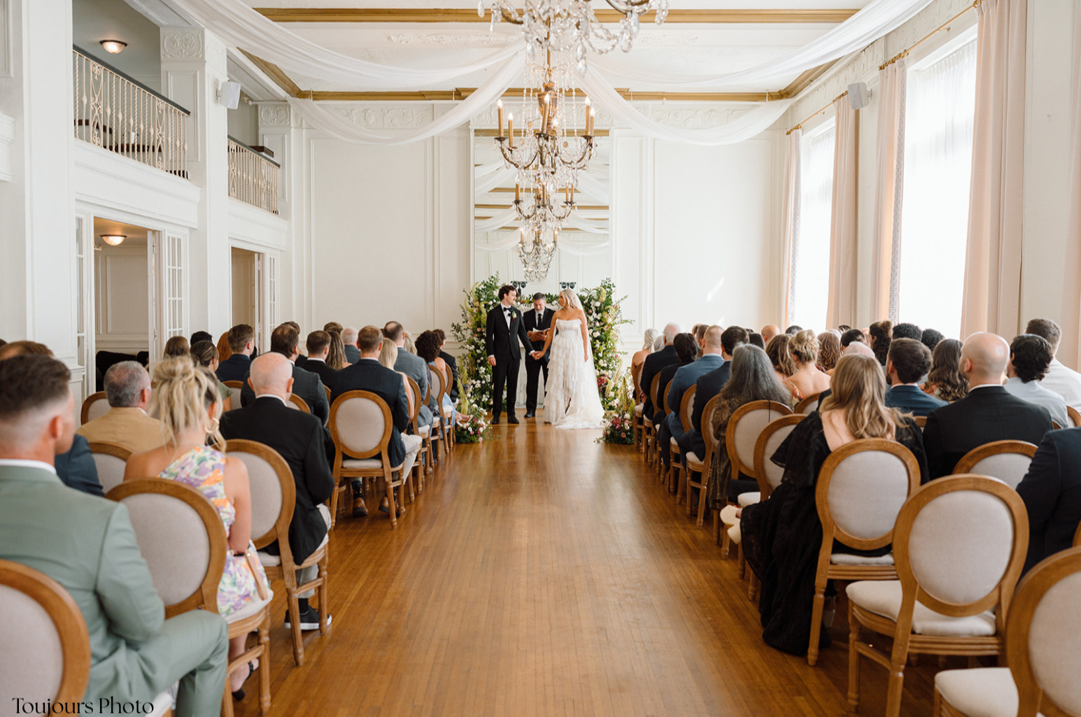 Bride and groom holding hands and standing before officiant during wedding ceremony in elegant, bright room decorated with white drapery, chandeliers, and floral arrangements.