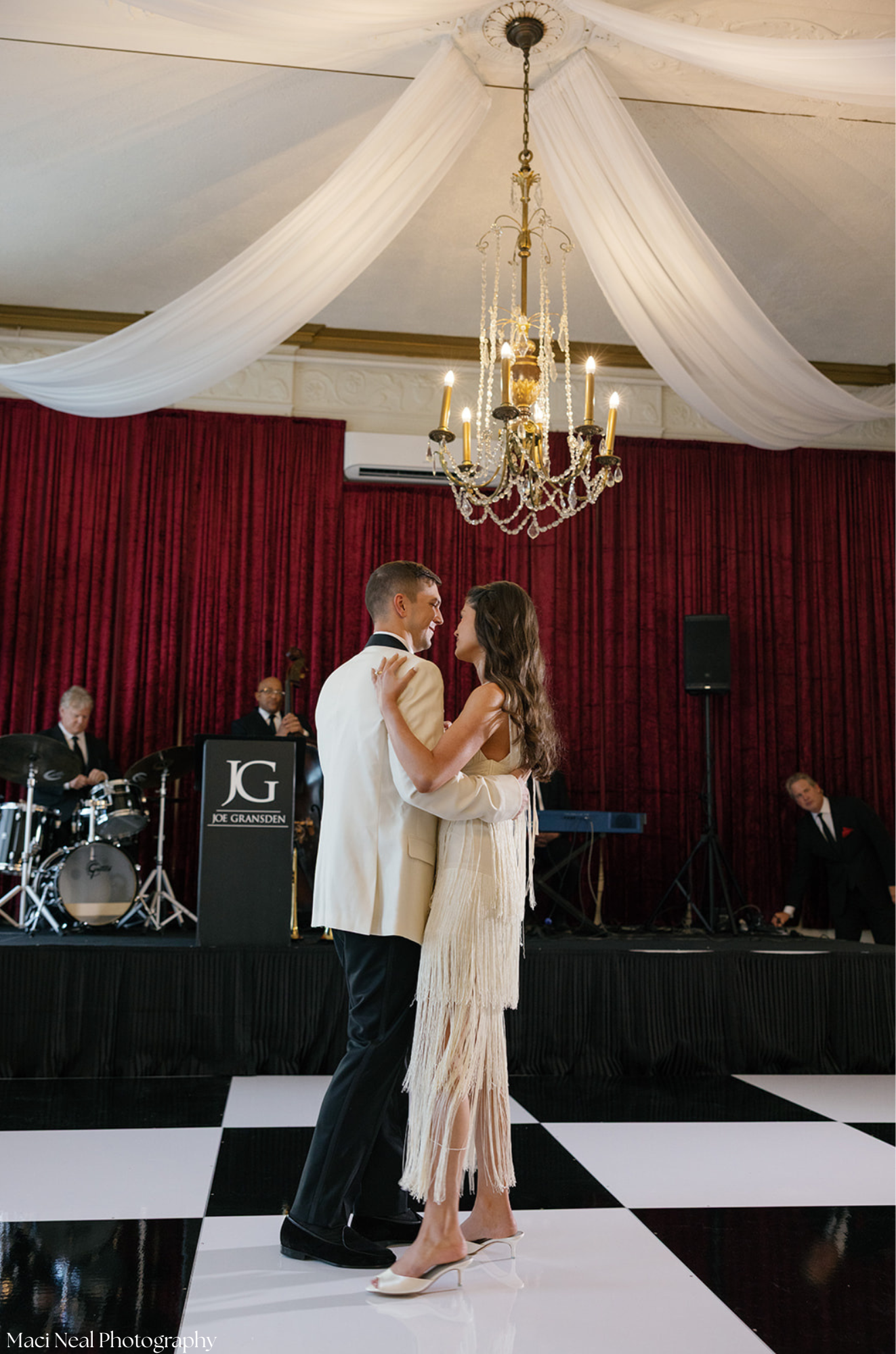 A couple is dancing on a checkered black and white dance floor, with a band playing music in the background, and red curtains behind them. The woman is wearing a fringed cream-colored dress and high heels, while the man is wearing a cream tuxedo jack