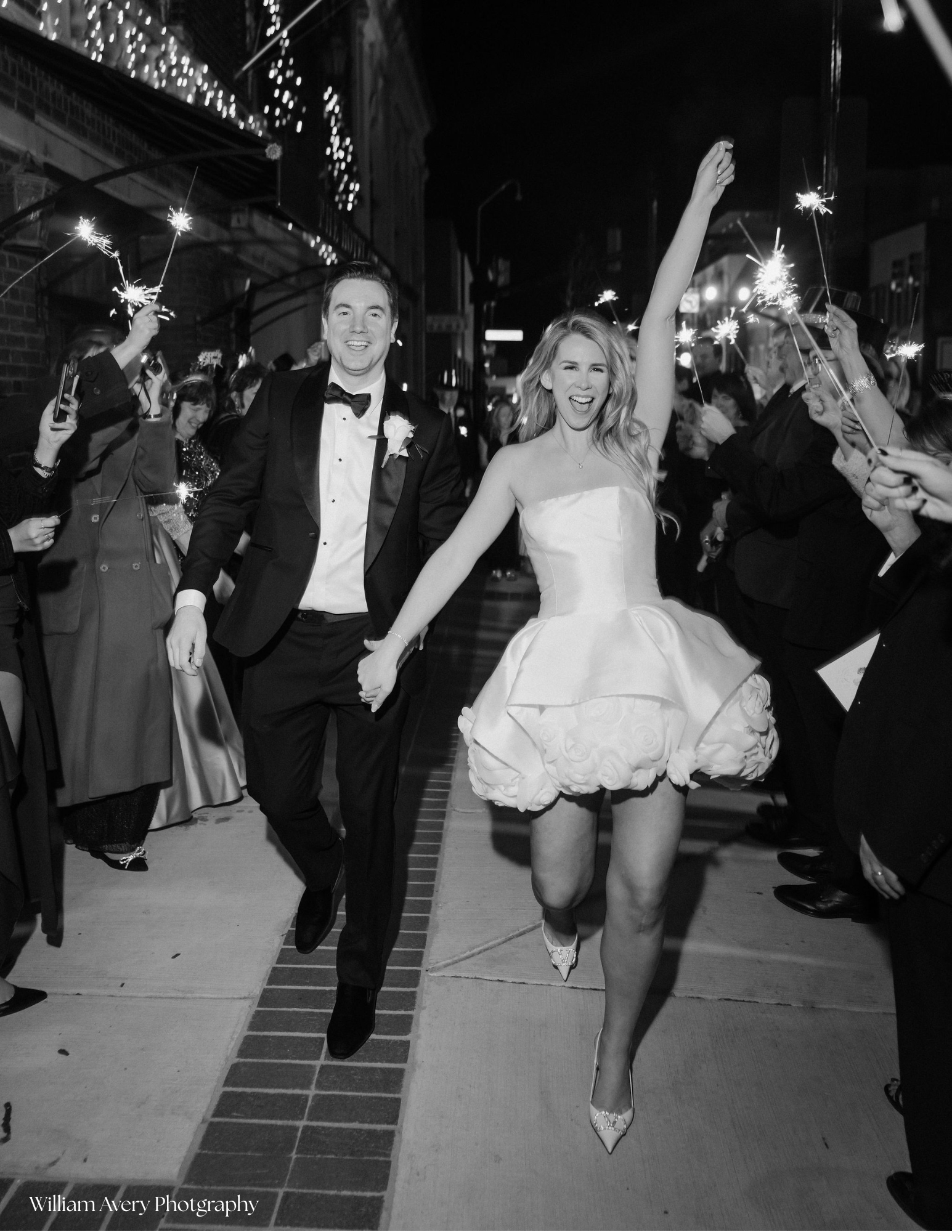 A bride and groom celebrating at night with sparklers as guests hold sparklers in a street decorated with hanging lights.