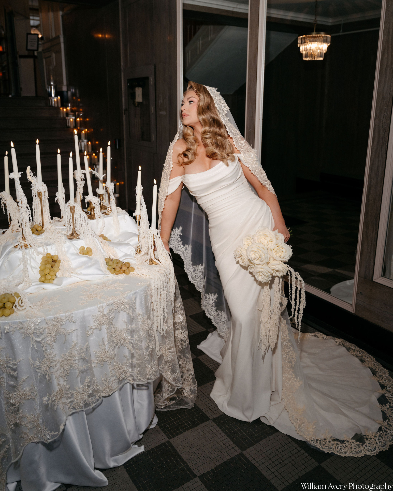 A bride in a white wedding dress with lace details and long train, holding a bouquet of white roses, is posing beside a table with lit candles and grapes, in a dimly lit room with dark walls and a chandelier.