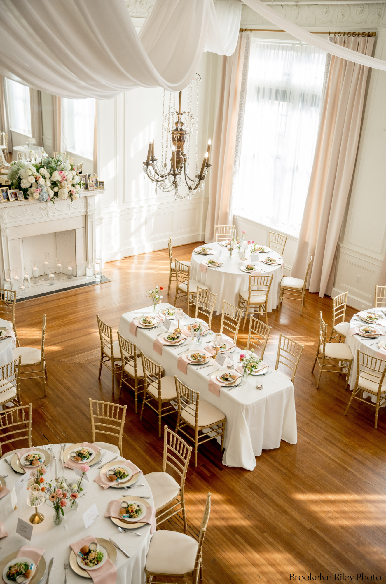 Elegant banquet hall decorated for a wedding or event with round tables set with floral centerpieces, place settings, and gold chairs, large windows with draped curtains, a chandelier, and a fireplace with floral arrangement on top.