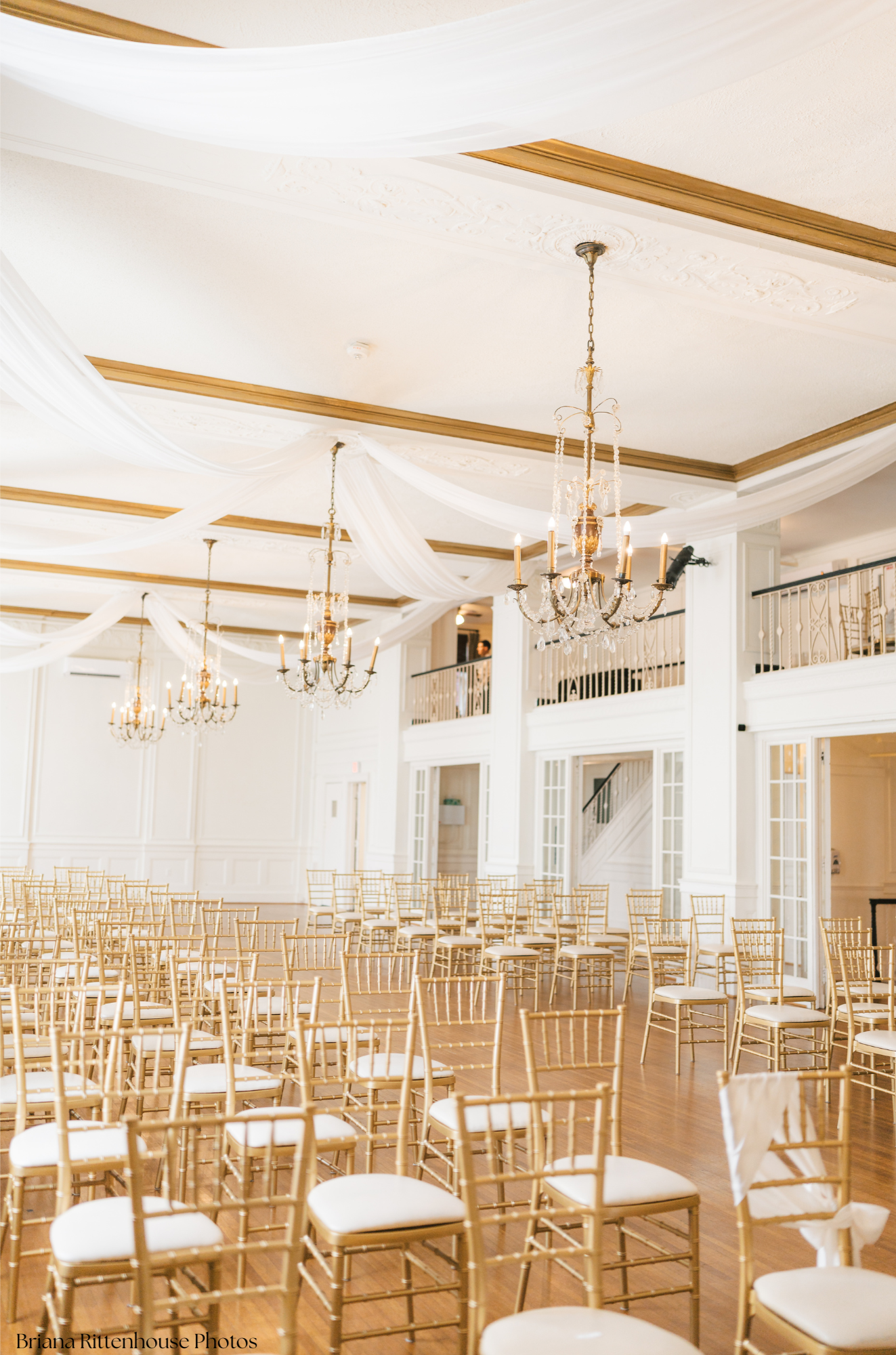 Elegant event space with rows of gold chairs with white cushions, crystal chandeliers hanging from a decorated white ceiling, draped white fabric, and large windows with white trim.