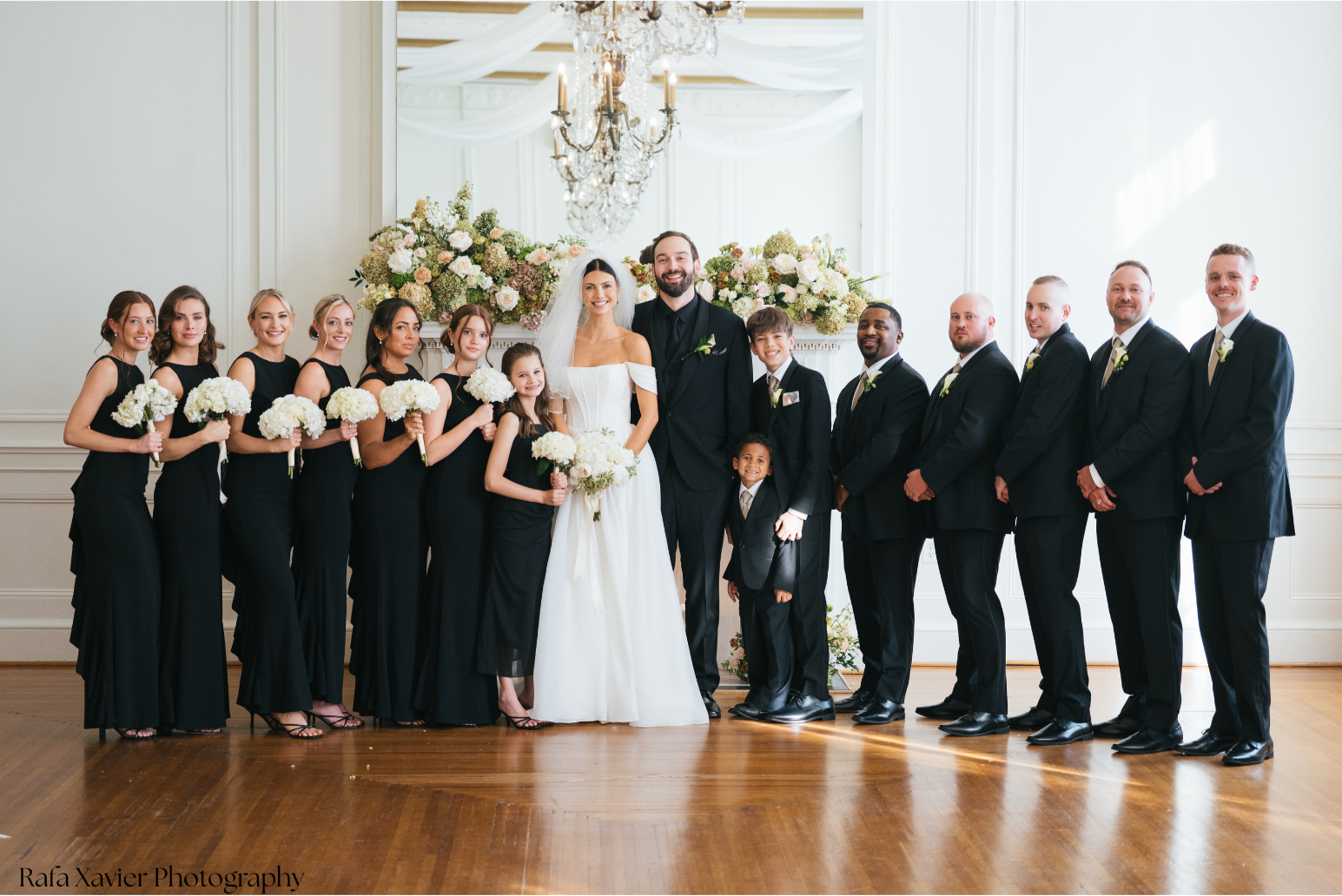A wedding party posing indoors with the bride and groom in the center, surrounded by bridesmaids and groomsmen, all smiling. The women are dressed in black dresses holding white bouquets, and the men are in black suits with boutonnieres. Two children