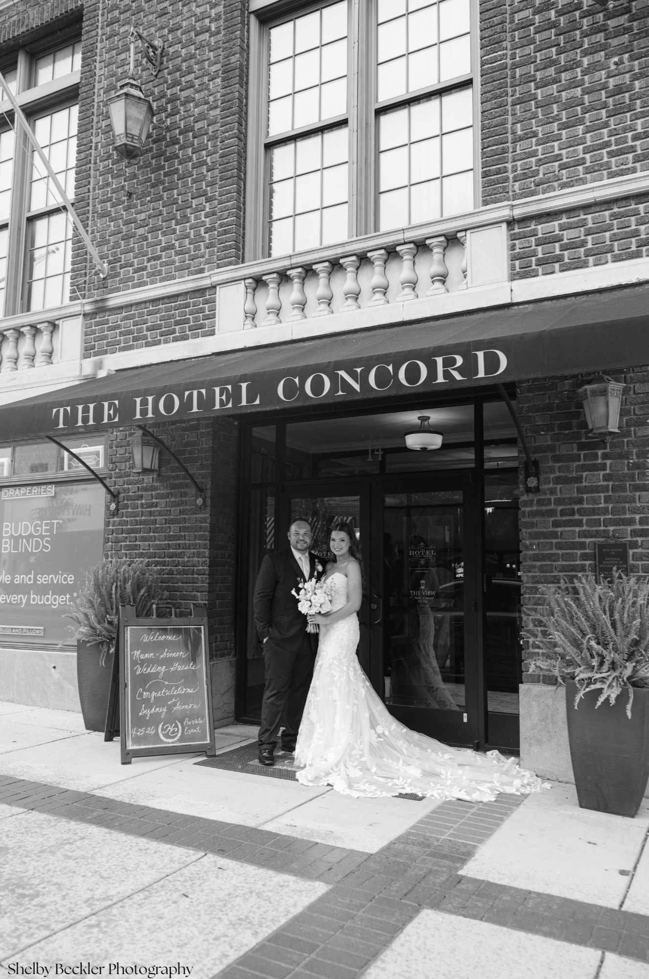 A bride and groom standing in front of a hotel entrance, celebrating their wedding. The bride is wearing a long lace wedding dress holding a bouquet, and the groom is in a dark suit. The hotel has a brick facade, a marquee with the words 'The Hotel C