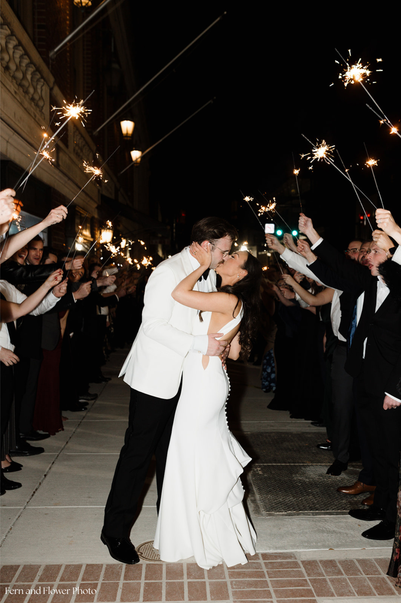 A newlywed couple kissing outside at night, surrounded by friends holding sparklers in celebration.