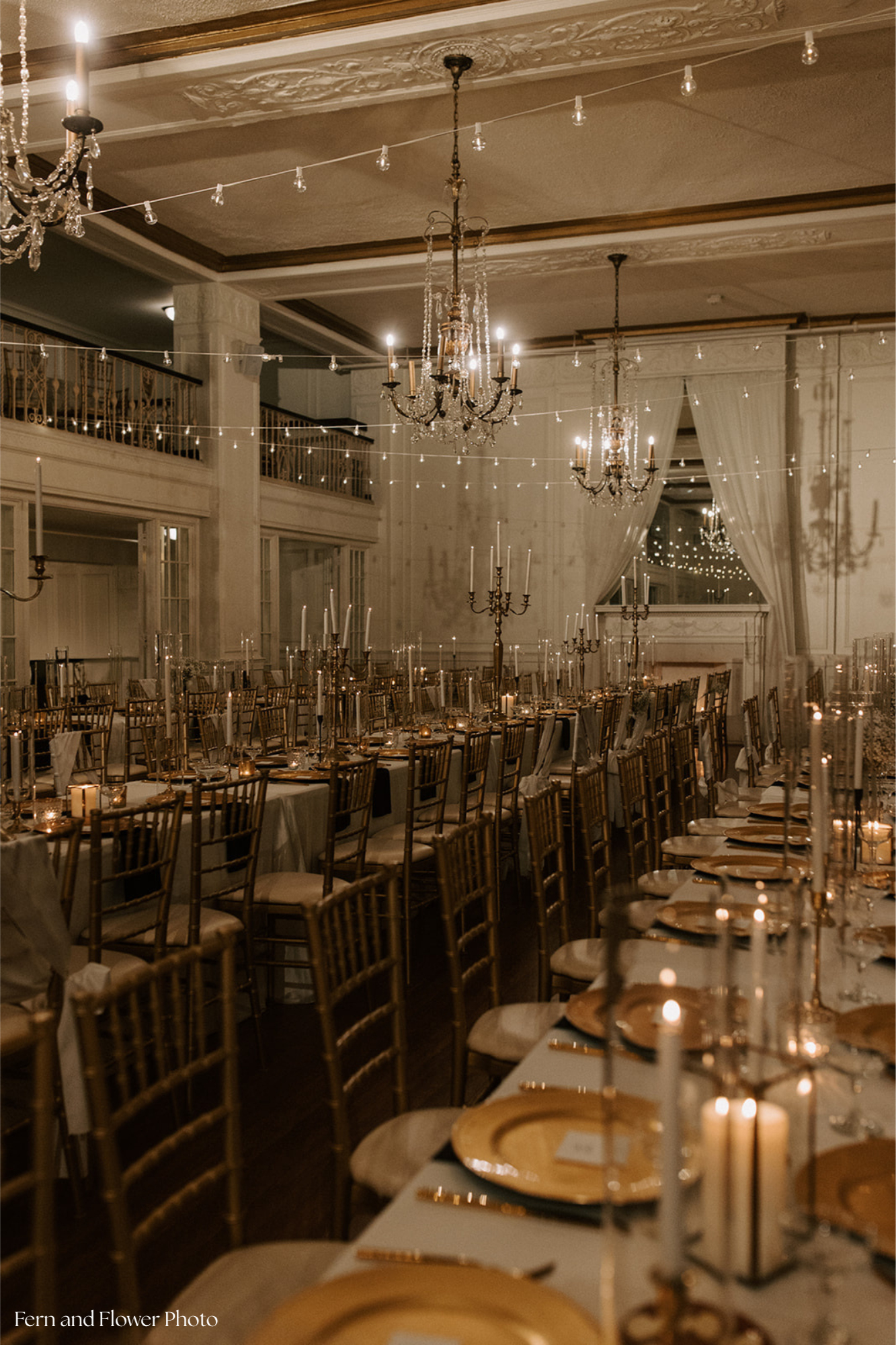 Elegant banquet hall decorated for a formal event with long tables, gold chairs, tall candelabras with candles, chandeliers, and string lights in a softly lit room.