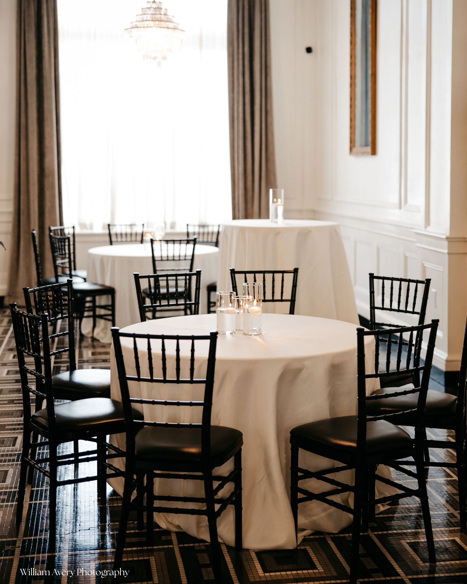 Elegant dining room with round tables covered in white tablecloths. Each table has a centerpiece of candles in glass holders. The room has large windows with curtains and classic wall paneling. The floor is patterned with black and beige tiles.