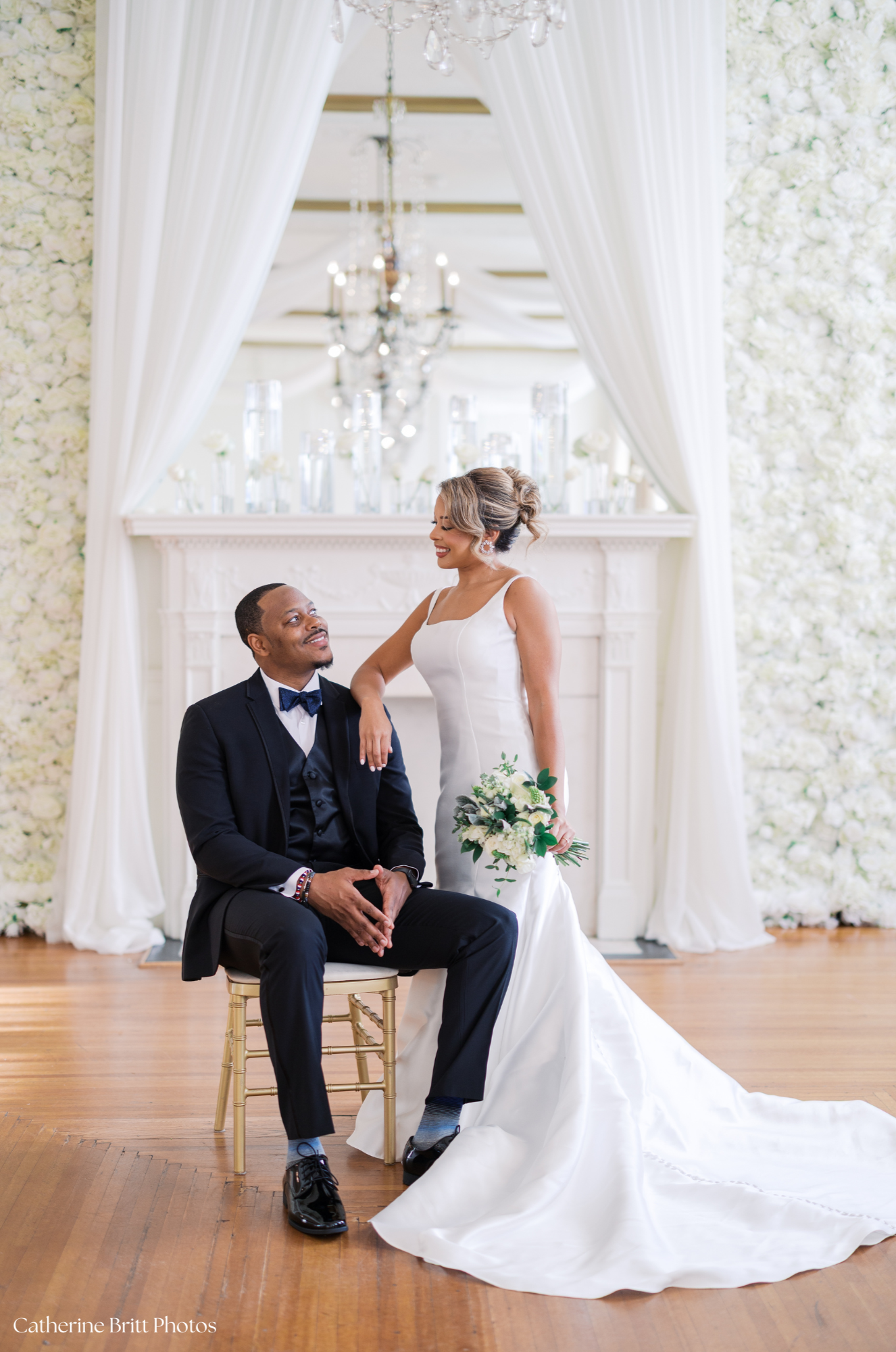 A bride and groom during their wedding ceremony in an elegant, bright room with white floral walls and draped curtains. The bride is standing with her hand on the groom's shoulder, smiling at him, while the groom is seated, looking up at her, smiling
