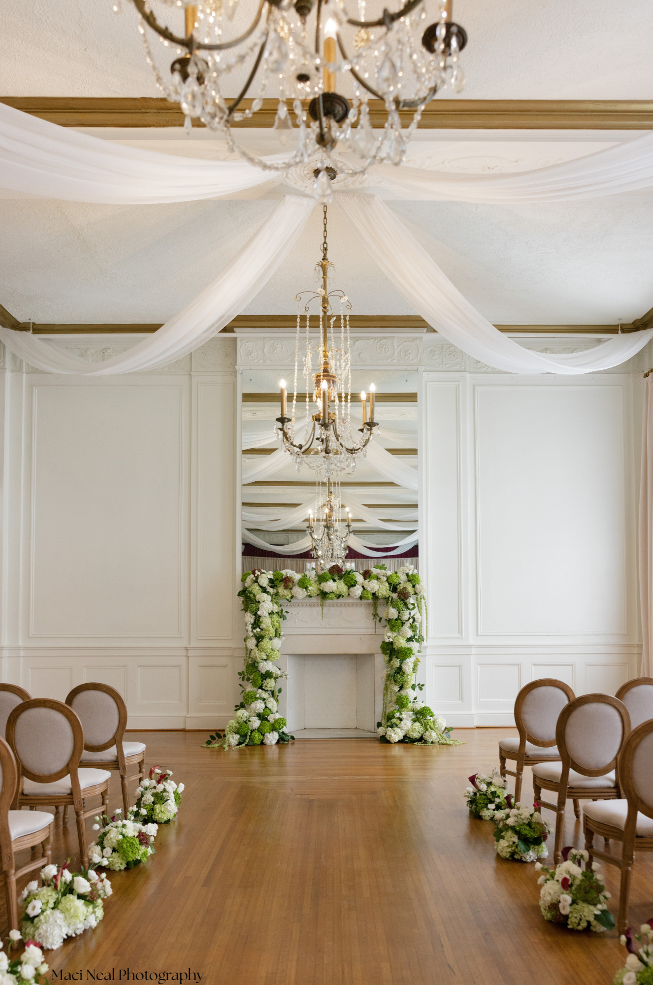 A decorated wedding ceremony venue with chandeliers, white drapes on the ceiling, a floral arch at the altar, and chairs arranged on either side of the aisle.