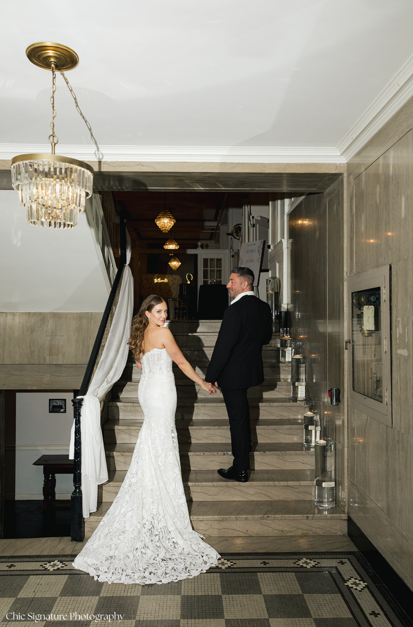 A bride and groom holding hands on a staircase inside a decorated venue, with elegant chandeliers overhead.