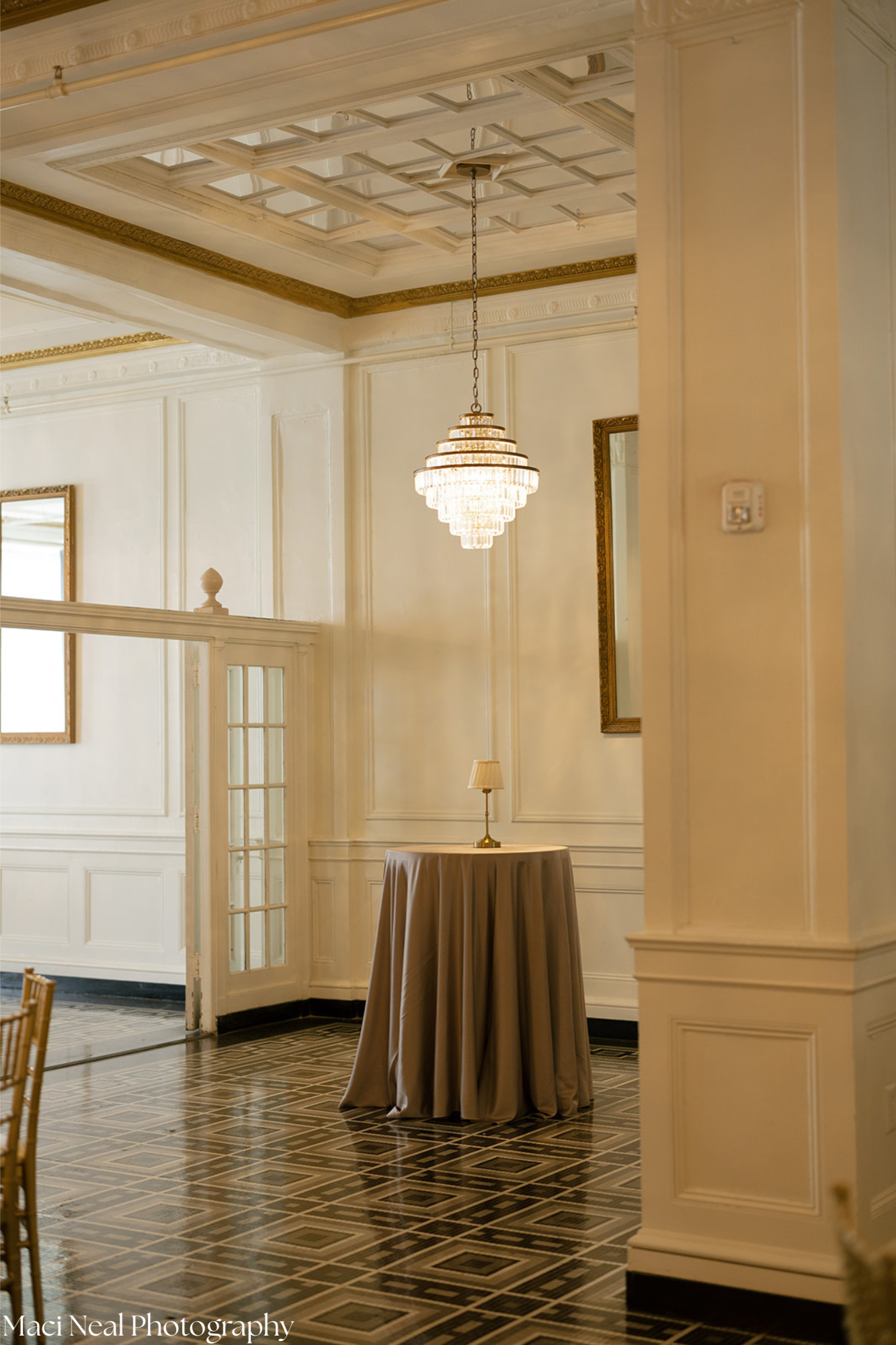 Interior of an elegant room with white paneled walls and ornate gold trim. A large chandelier hangs from the ceiling, illuminating the space. There is a tall, round table covered with a beige tablecloth, and a small table lamp is placed on top. Frame