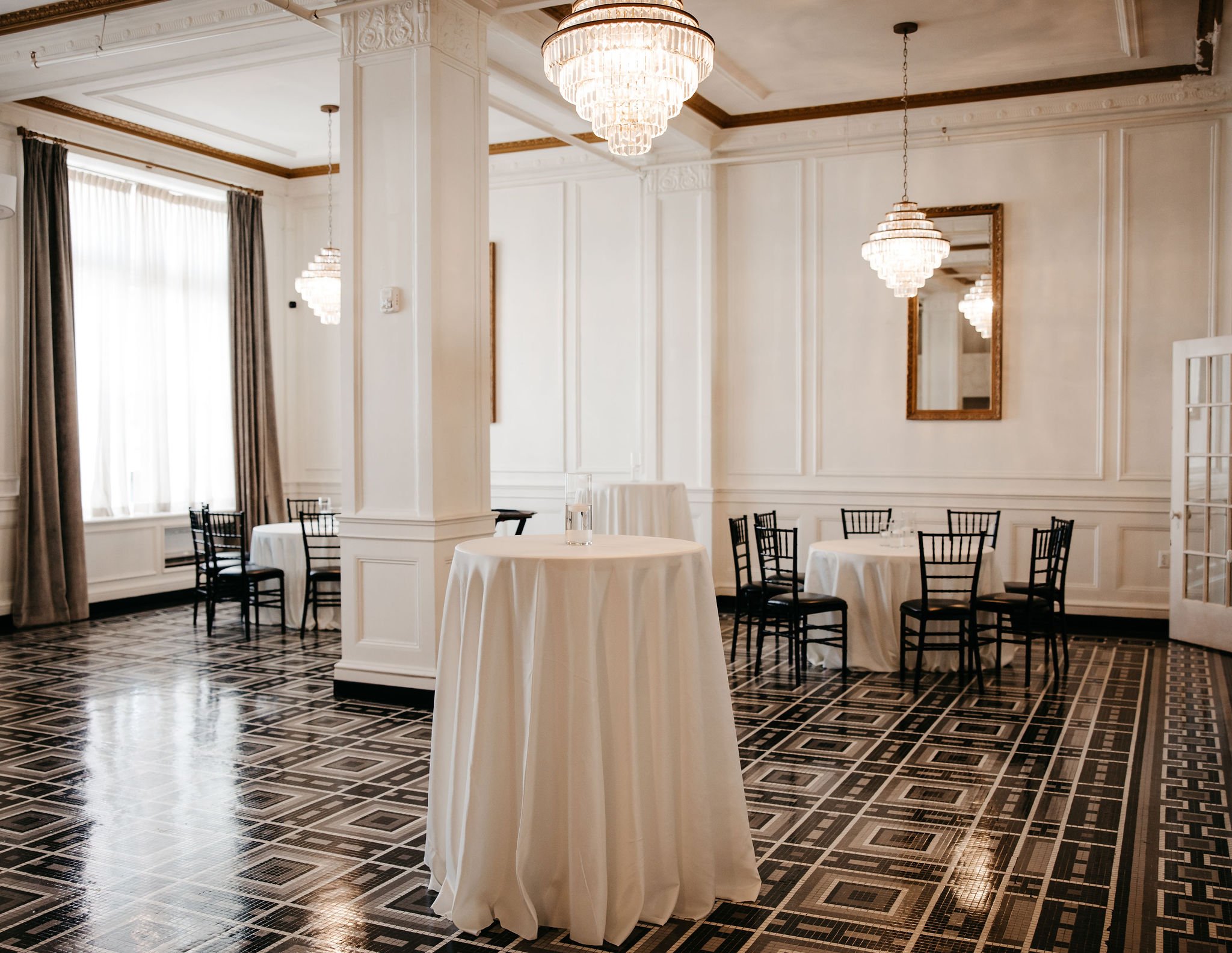 Elegant banquet hall decorated with white walls, ornate moldings, large chandelier lighting fixtures, tall windows with curtains, and round tables with white tablecloths and black chairs.