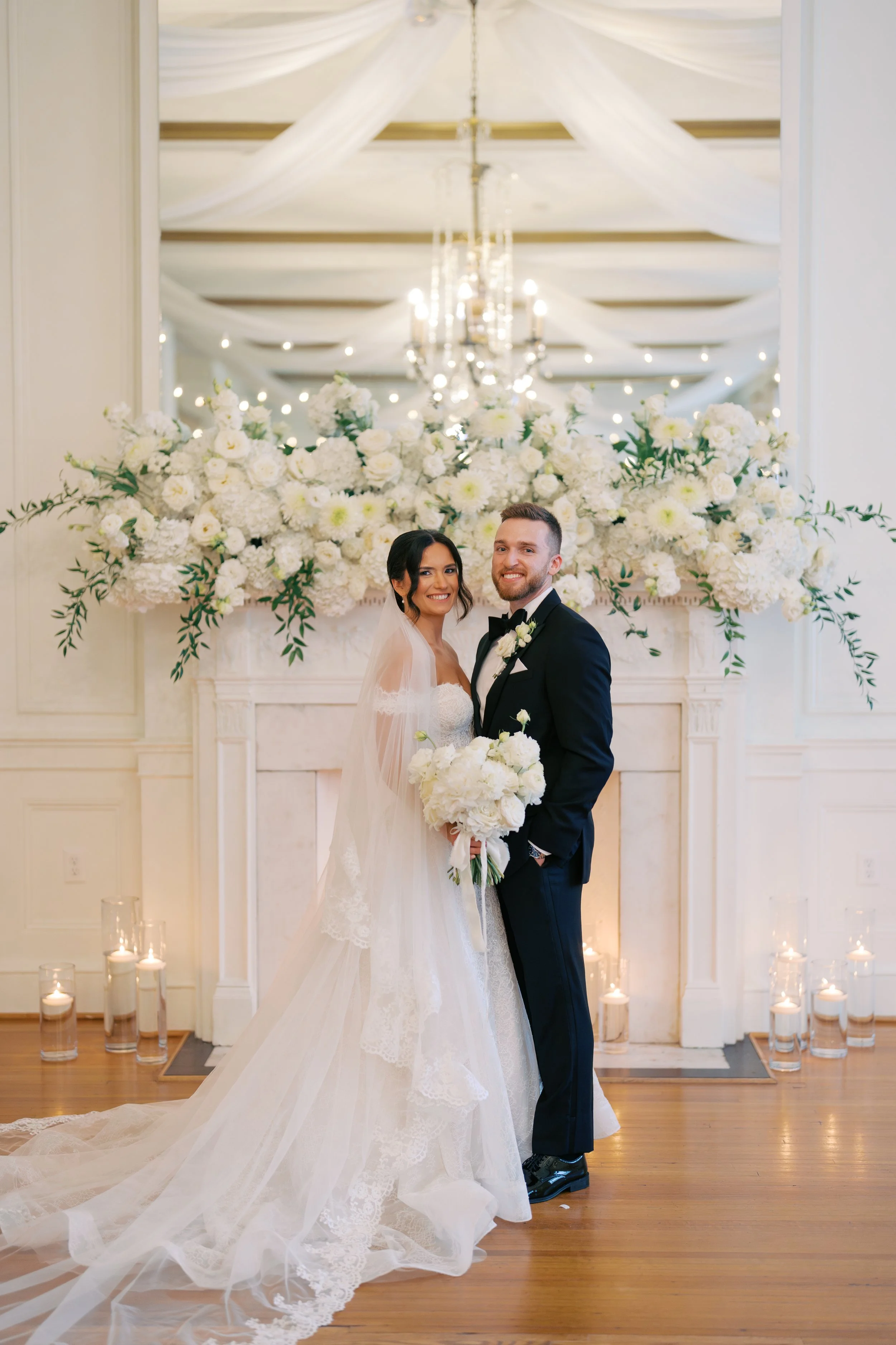 A bride and groom standing together at their wedding ceremony in front of a large floral arrangement with white flowers and greenery, an ornate mirror, and wedding candles on the floor. The bride is wearing a white lace wedding dress and veil, holdin
