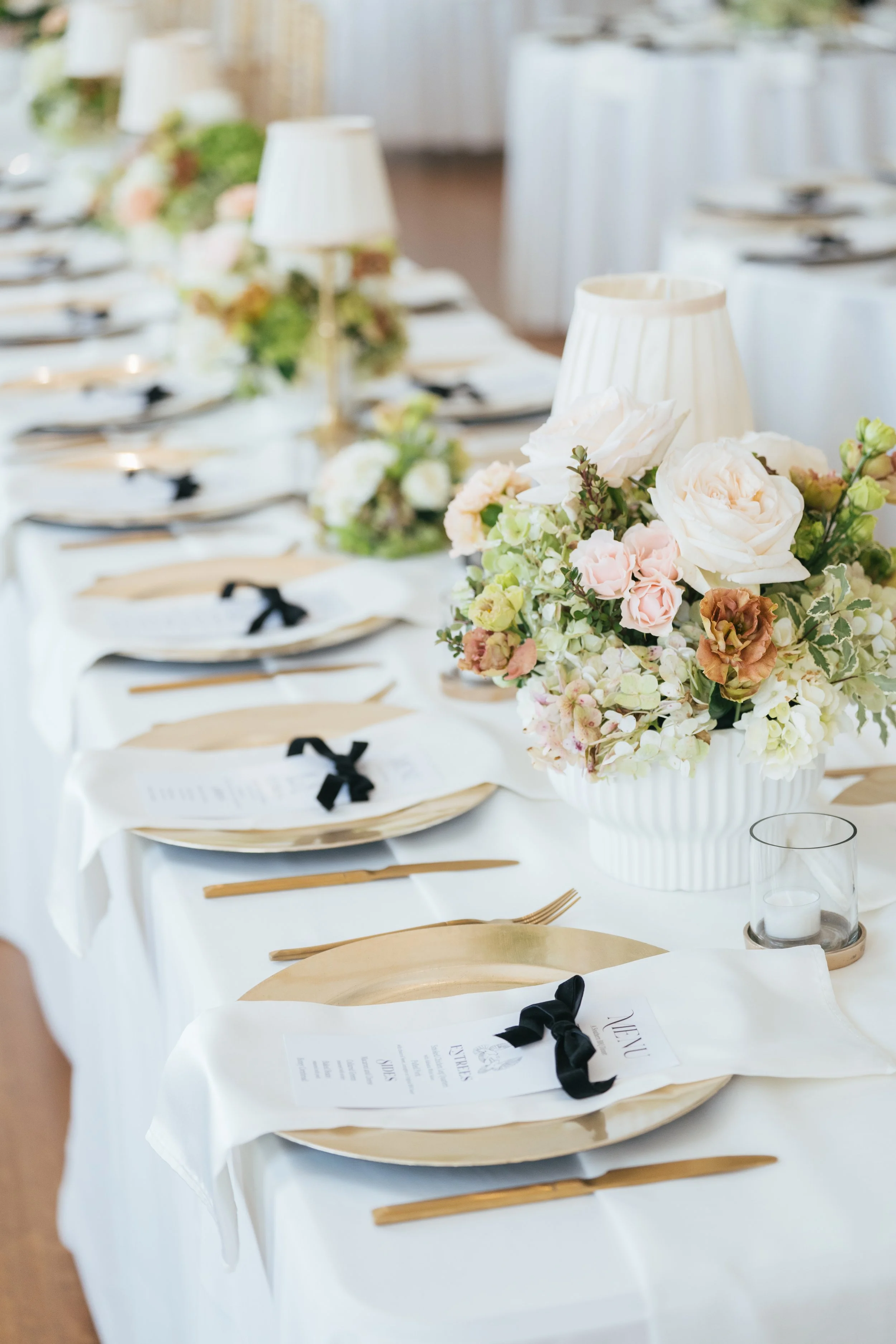 A decorated banquet table with floral centerpieces, white lamps, candles, and place settings including menus and cutlery.