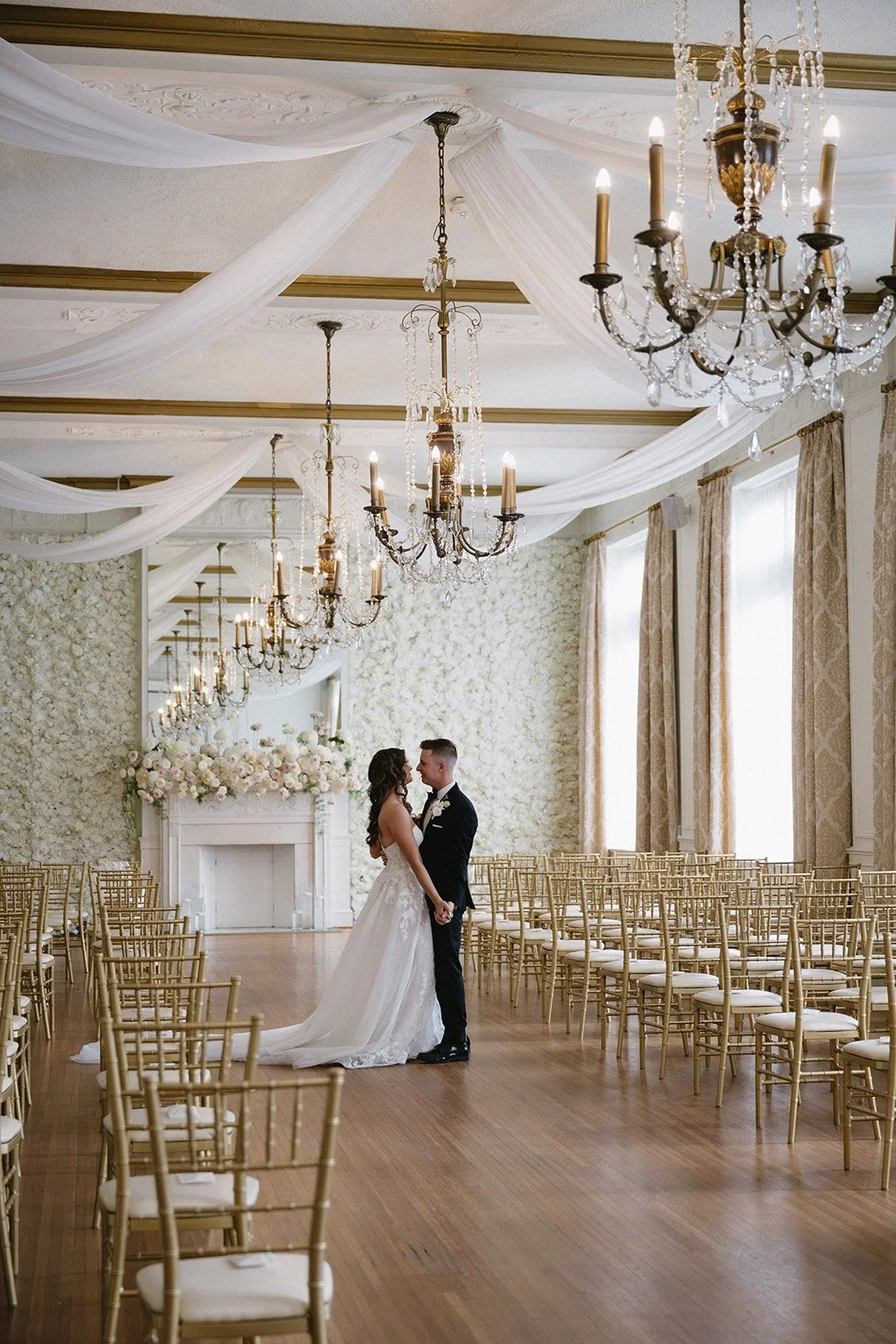 A bride and groom holding hands and gazing at each other in a decorated wedding venue with chandeliers, draped fabric on the ceiling, a floral fireplace mantle, and rows of gold chairs.