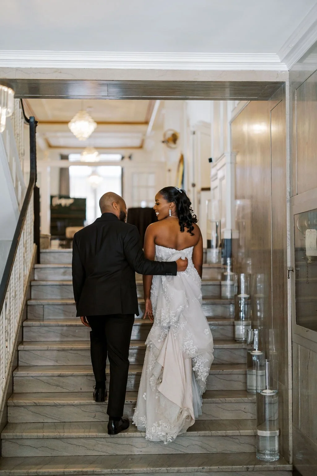 A bride and groom walking up a marble staircase in an elegant, well-lit building, smiling at each other after their wedding ceremony.