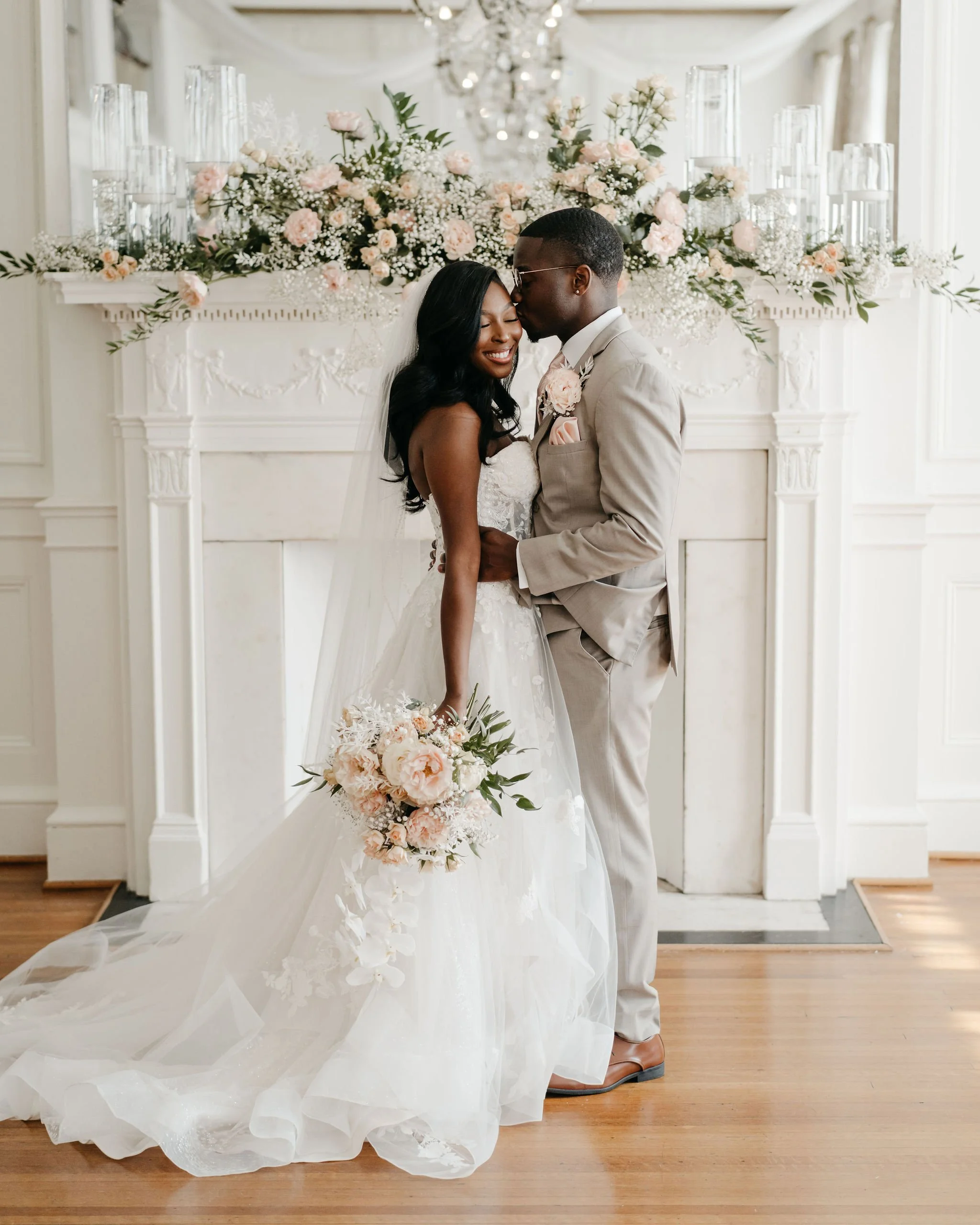 A bride and groom sharing a kiss in front of a decorated fireplace with pink and white flowers and candles, at their wedding celebration.