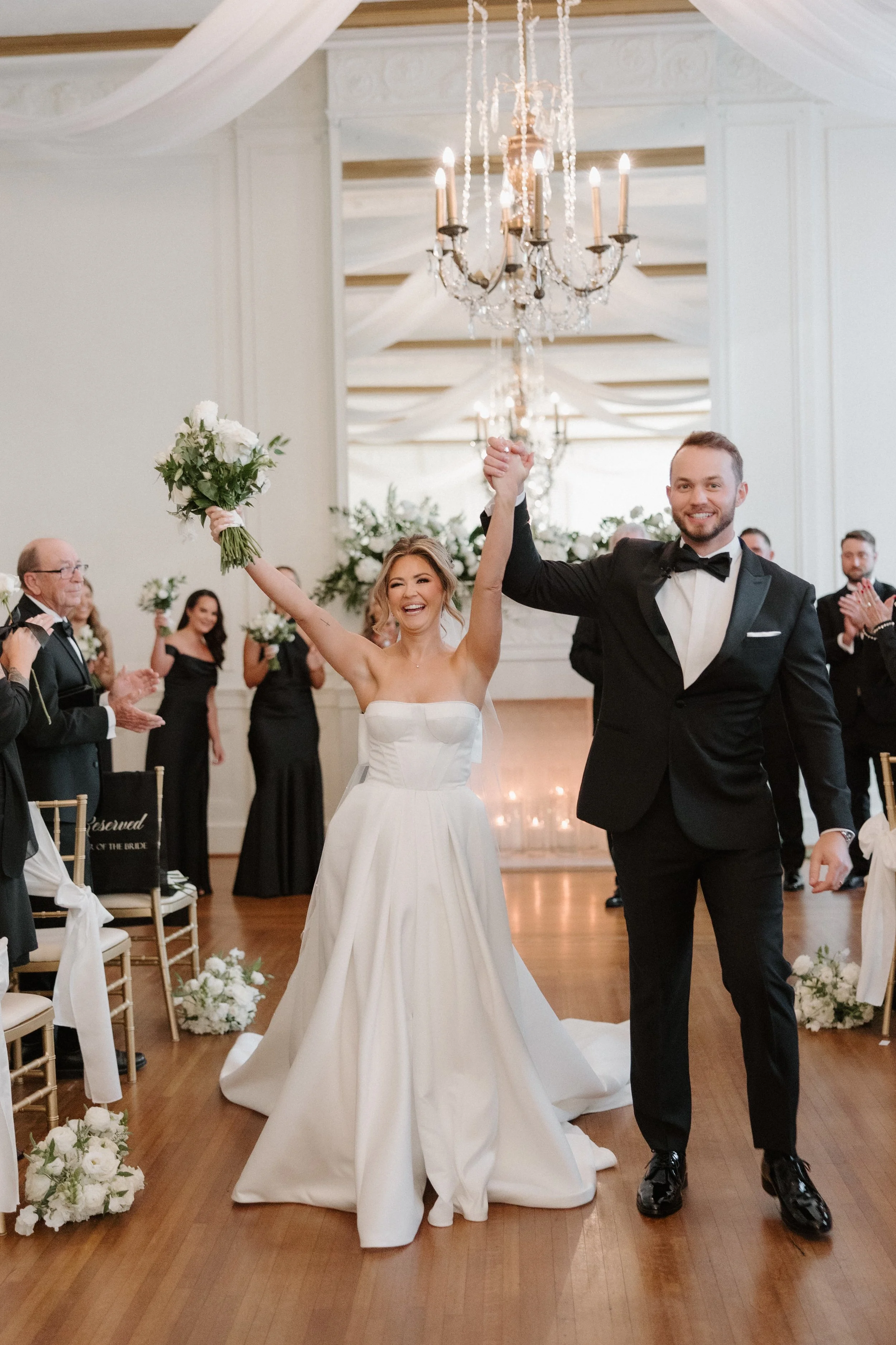 A bride and groom celebrating their wedding ceremony indoors, smiling and holding hands up. The bride is in a white wedding dress holding a bouquet, and the groom is in a black tuxedo. Guests are clapping in the background, and the room is decorated 
