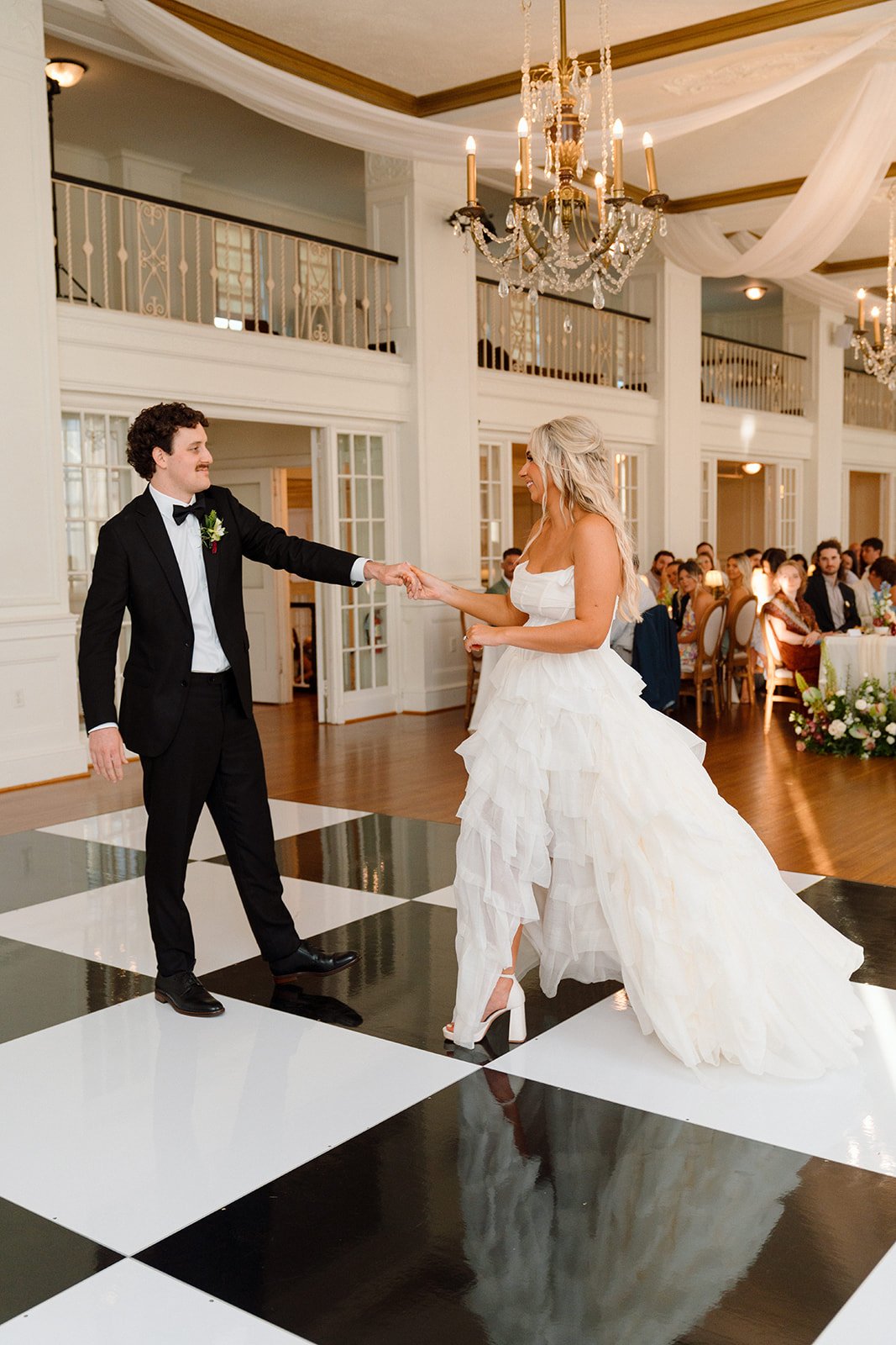 Bride and groom dancing at their wedding reception in a grand hall with chandeliers, white drapery, and guests seated at tables.
