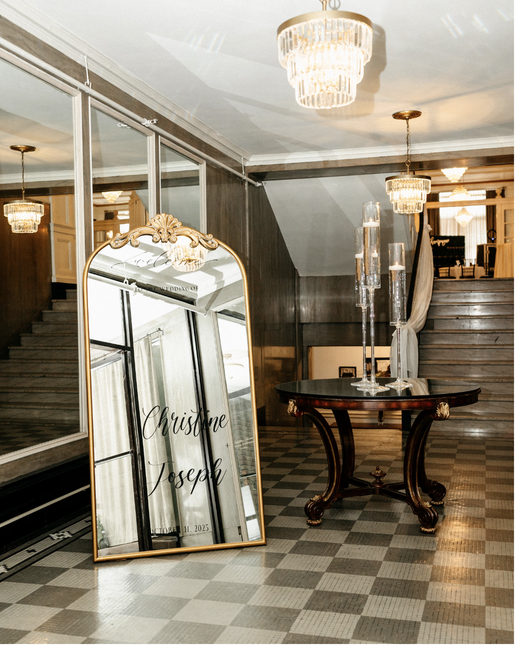 Elegant hotel lobby with a tall, ornate mirror with gold accents, a dark wood table with glass candle holders, and a staircase in the background; chandelier and wall sconces providing warm lighting.
