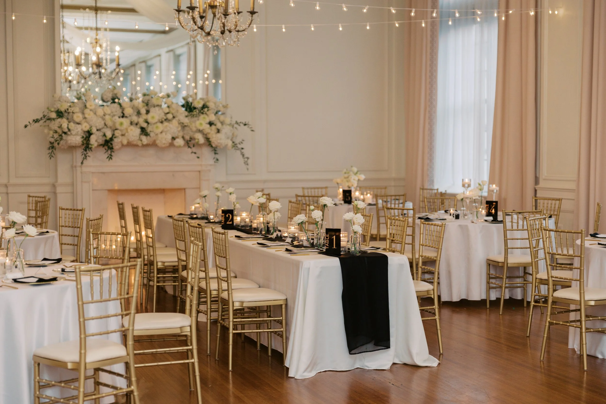 Elegant banquet hall decorated for a wedding or formal event with tables set with white tablecloths, black napkins, white flower centerpieces, and gold chairs. A large floral arrangement and chandeliers are visible on the wall behind.