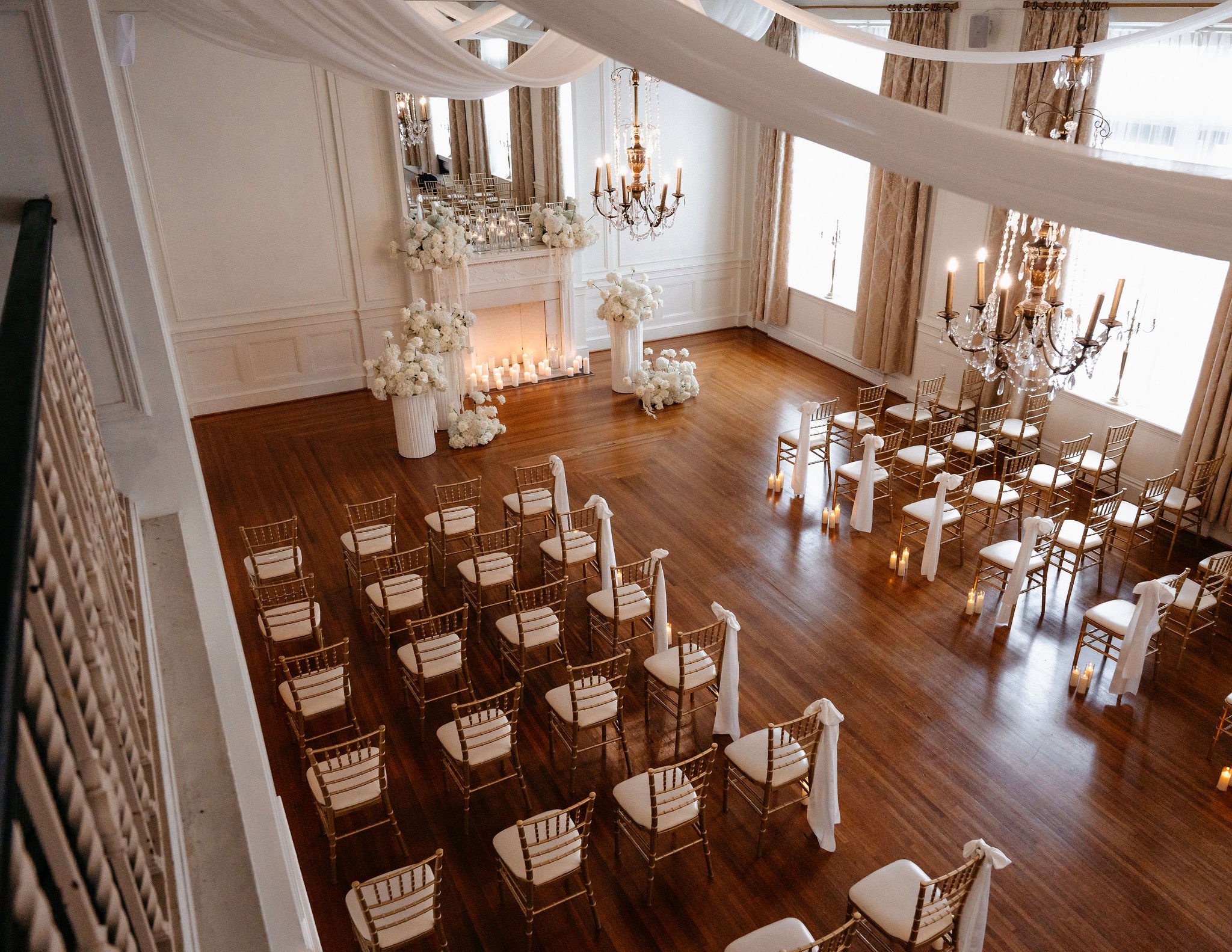 Elegant wedding ceremony setup in a spacious room with a decorated fireplace, white flowers, candles, chandeliers, gold chairs, and draped fabric.