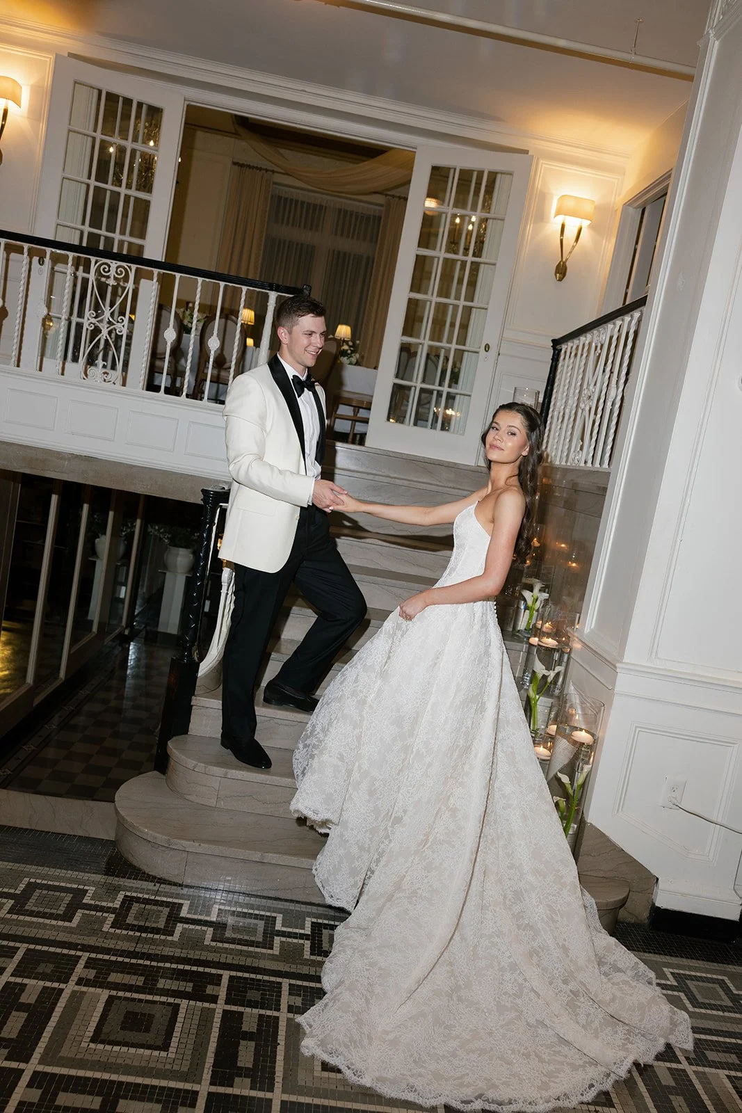 A newlywed couple dancing on a staircase in an elegant venue, with the groom in a tuxedo and the bride in a lace wedding gown.