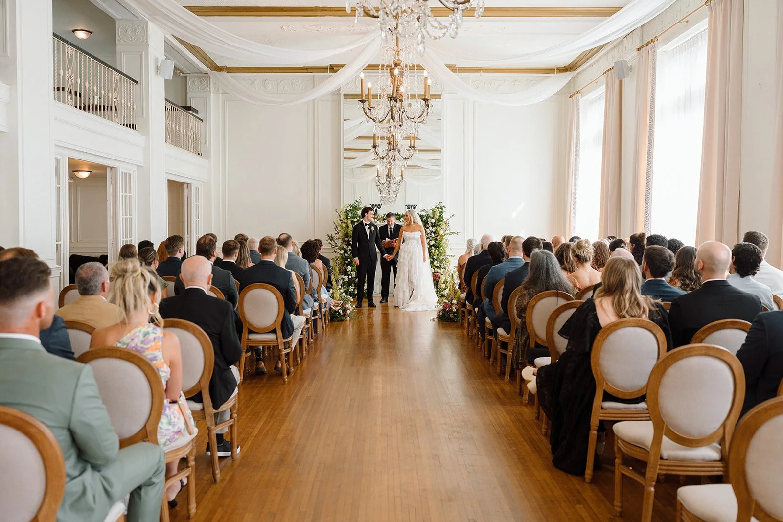 A wedding ceremony in a bright, elegant room with white walls and large windows. The bride and groom are standing at the altar, holding hands, with an officiant in between. Guests are seated on both sides, facing the couple. The room features chandel